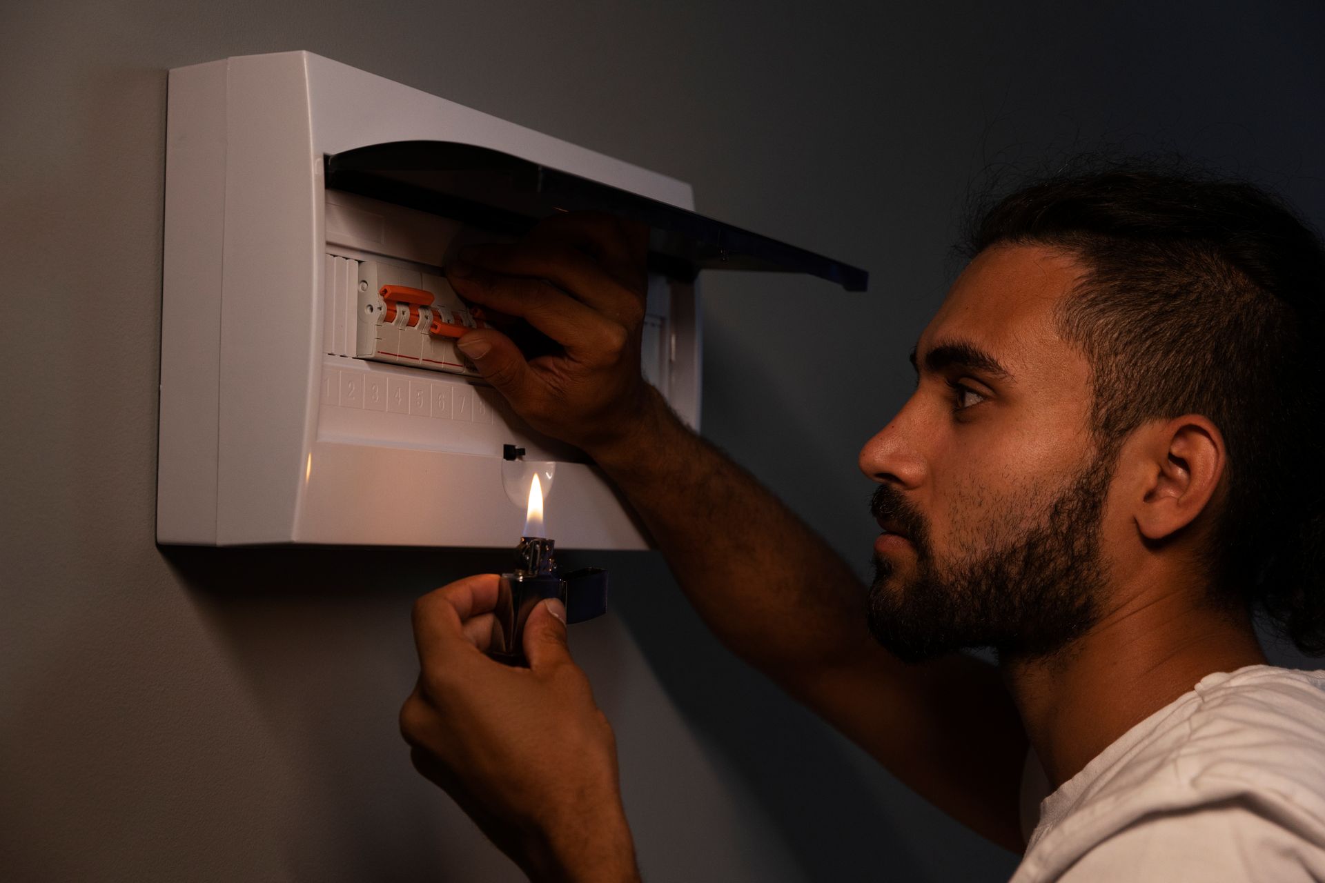 A man using a lighter to see inside an open electrical panel on a gray wall.