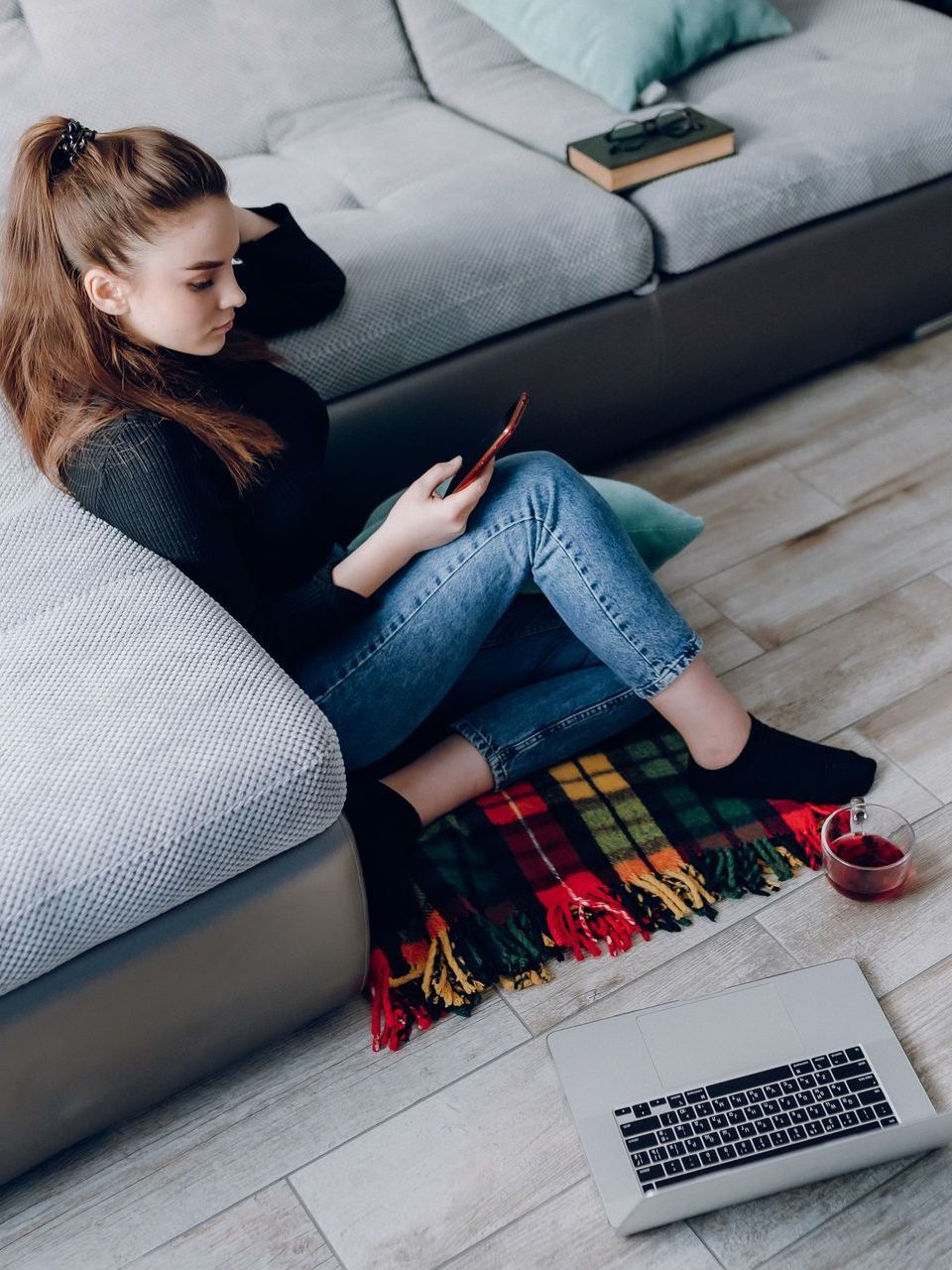 a woman sitting on the floor, holding her phone and her laptop is on the floor along with a cup of tea