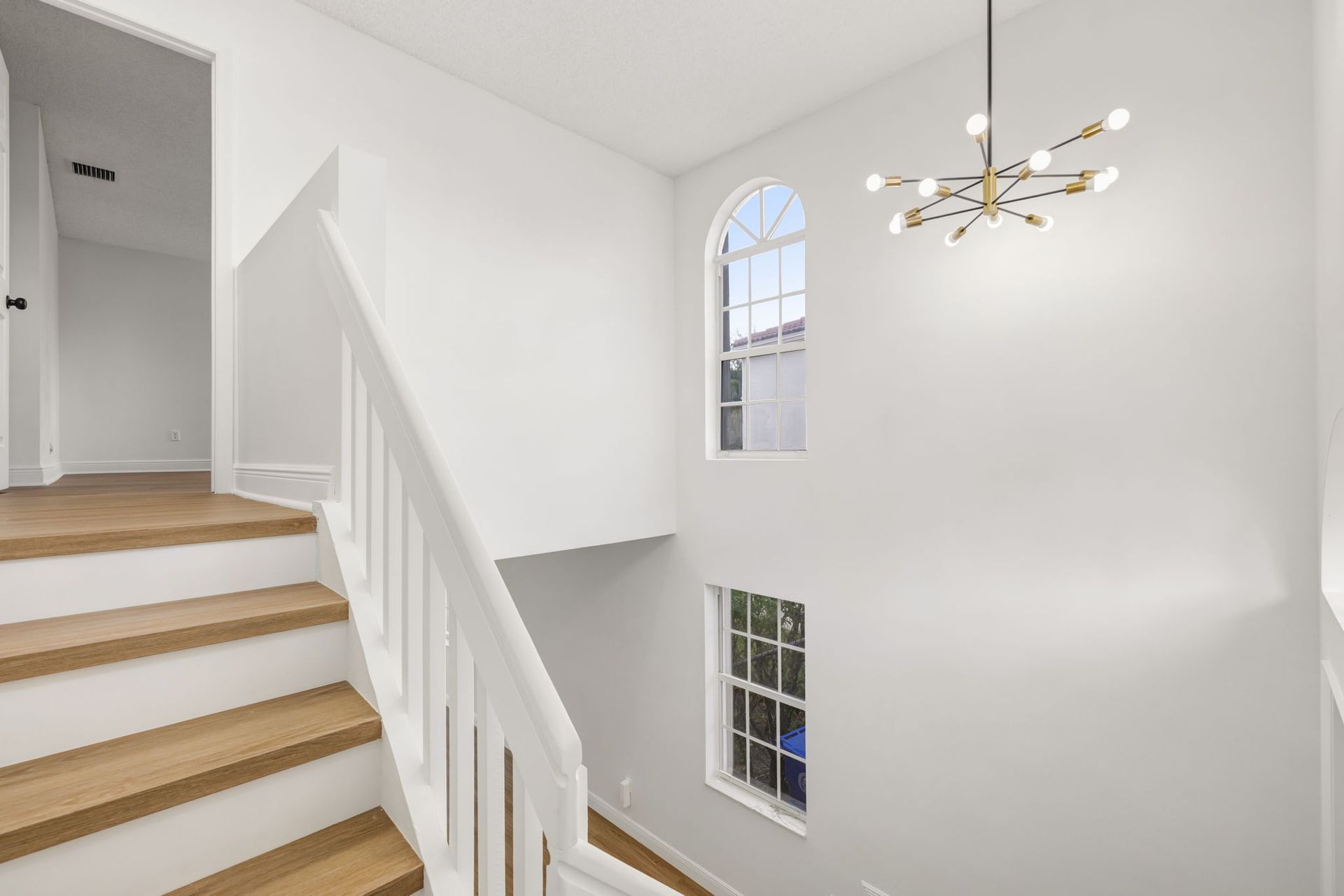 A modern staircase with light wood steps and white railings leads upward next to two windows and a starburst chandelier.