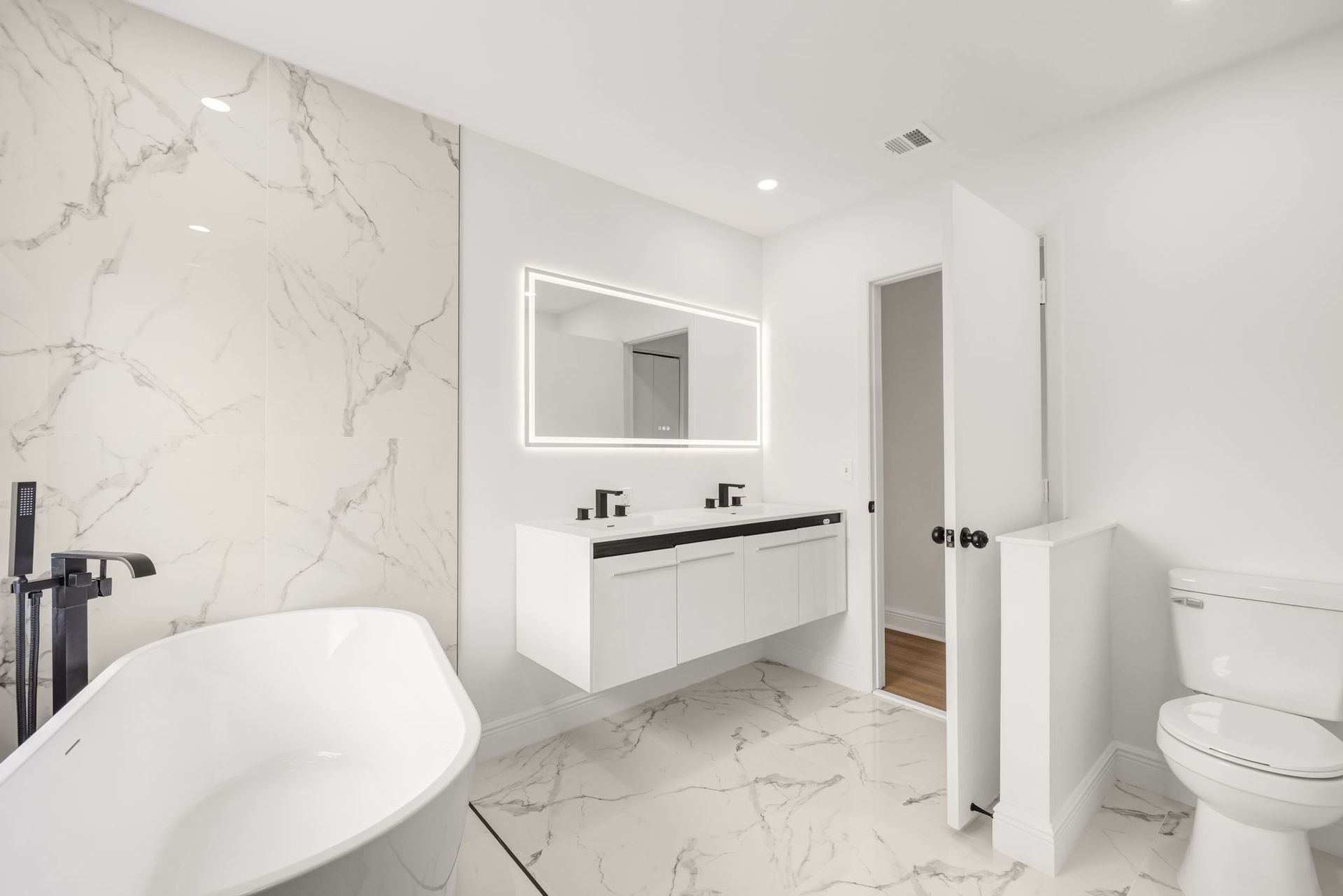A modern bathroom featuring a white freestanding tub, a floating vanity with a lit mirror, and white marble-patterned tiles.