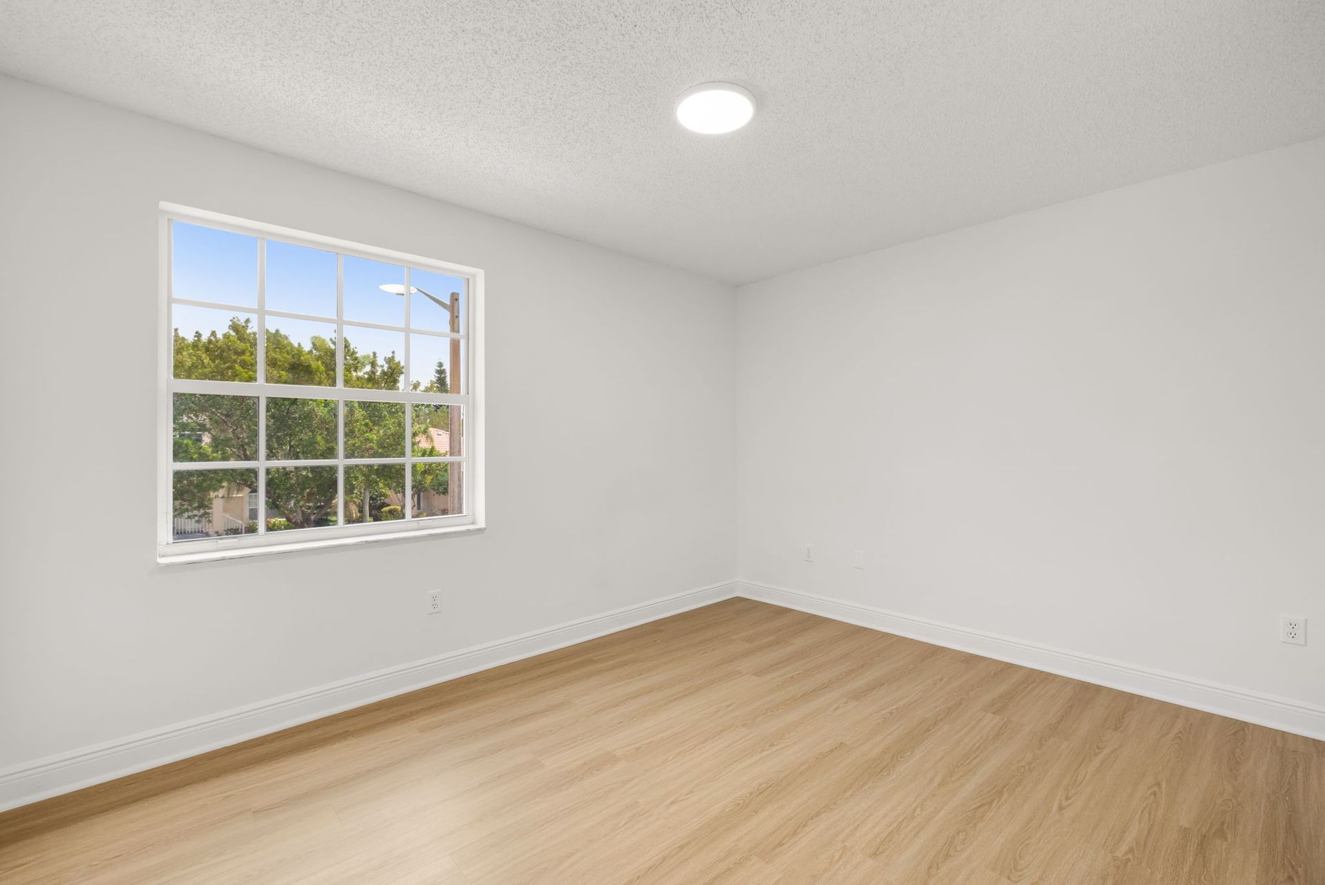 An empty room with white walls, a single window looking out at trees, and light-colored wood flooring under a ceiling light.