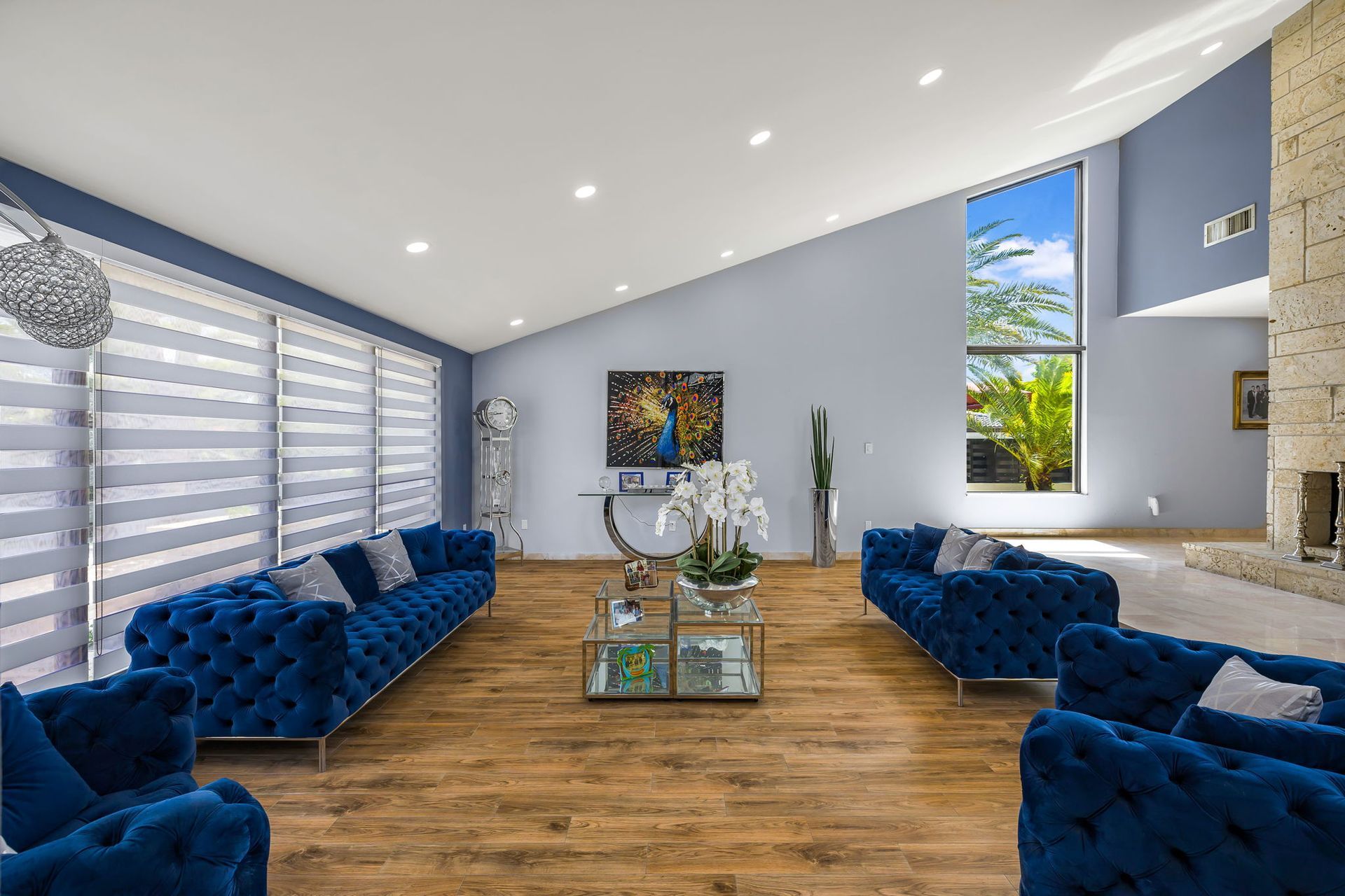A spacious, modern living room with blue velvet couches, wooden floors, and large windows.