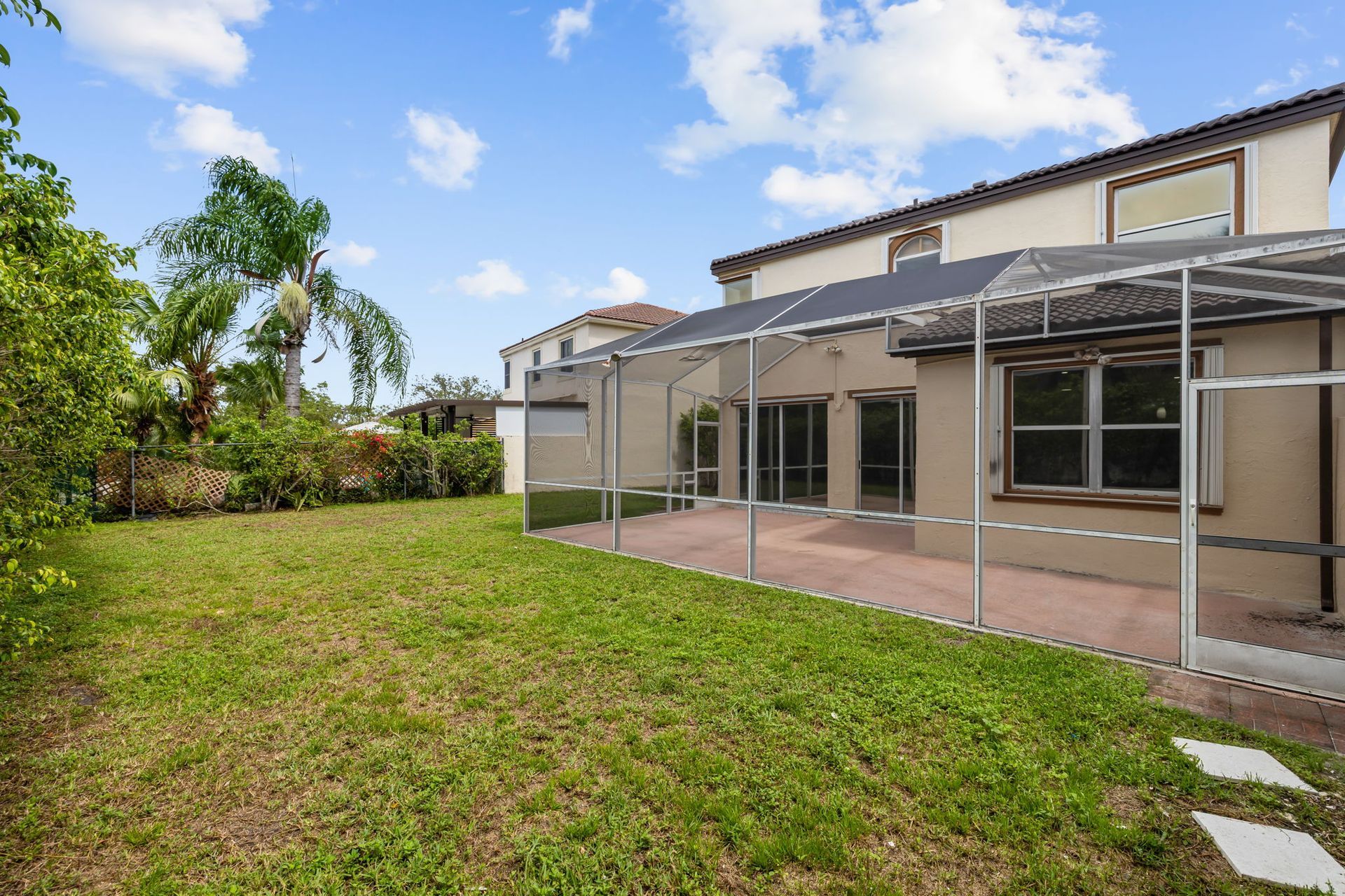 A green grassy lawn in the backyard of a two-story beige house with a screened-in patio under a bright, cloudy blue sky.