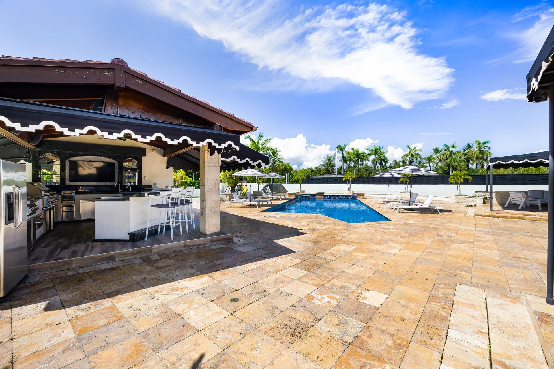Patio with outdoor kitchen, pool, and lounge chairs under a bright blue sky.