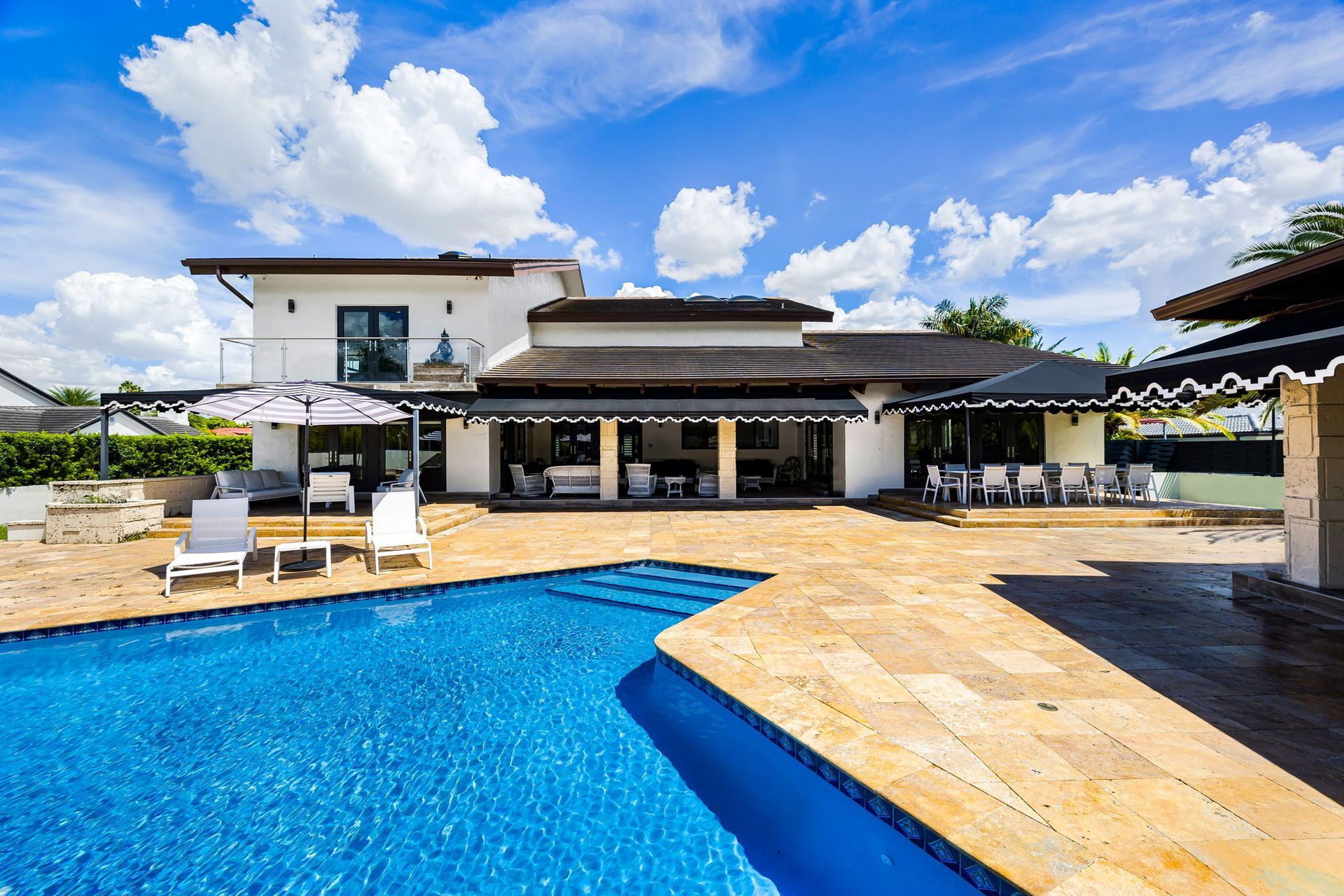 Patio with outdoor kitchen, swimming pool, and lounge chairs under a bright blue sky.