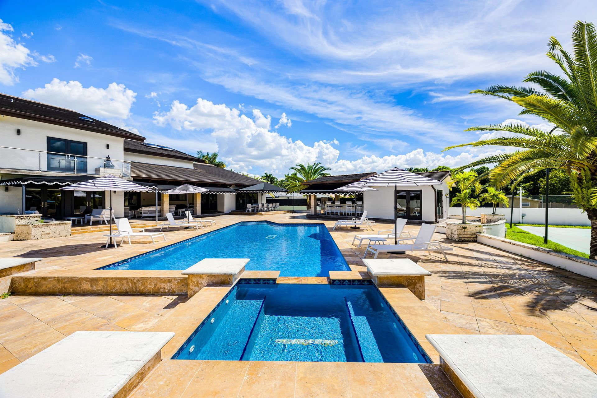 Luxurious outdoor pool area with a house, blue pool, lounge chairs, and sunny sky.
