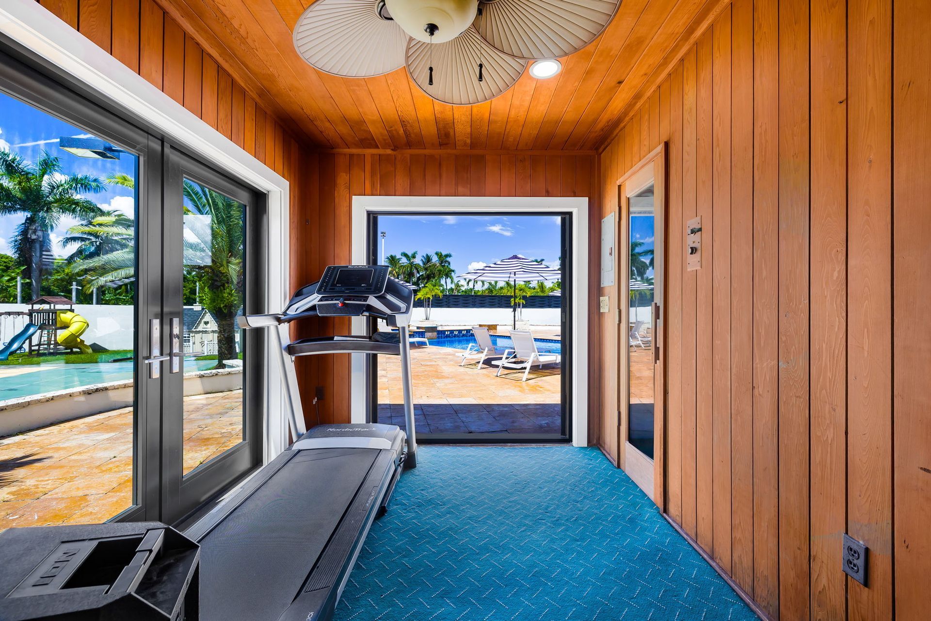 Treadmill in a small room with a pool view, blue carpet, and wood paneling.