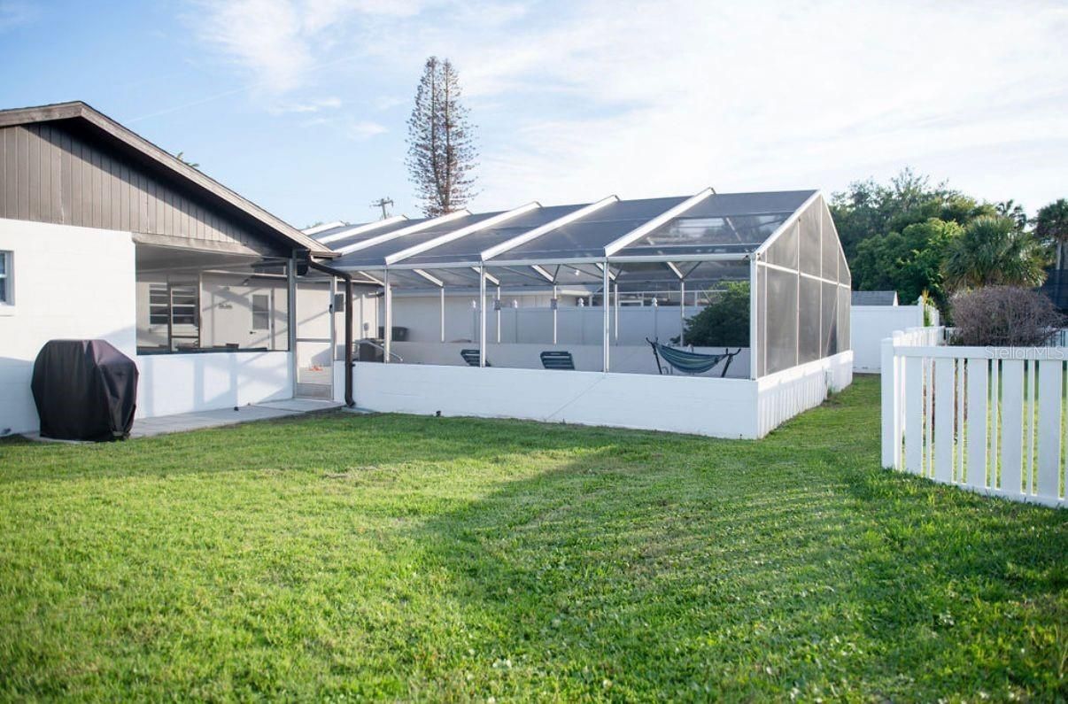 Patio trasero con terraza blanca cerrada y césped junto a una casa bajo un cielo soleado.