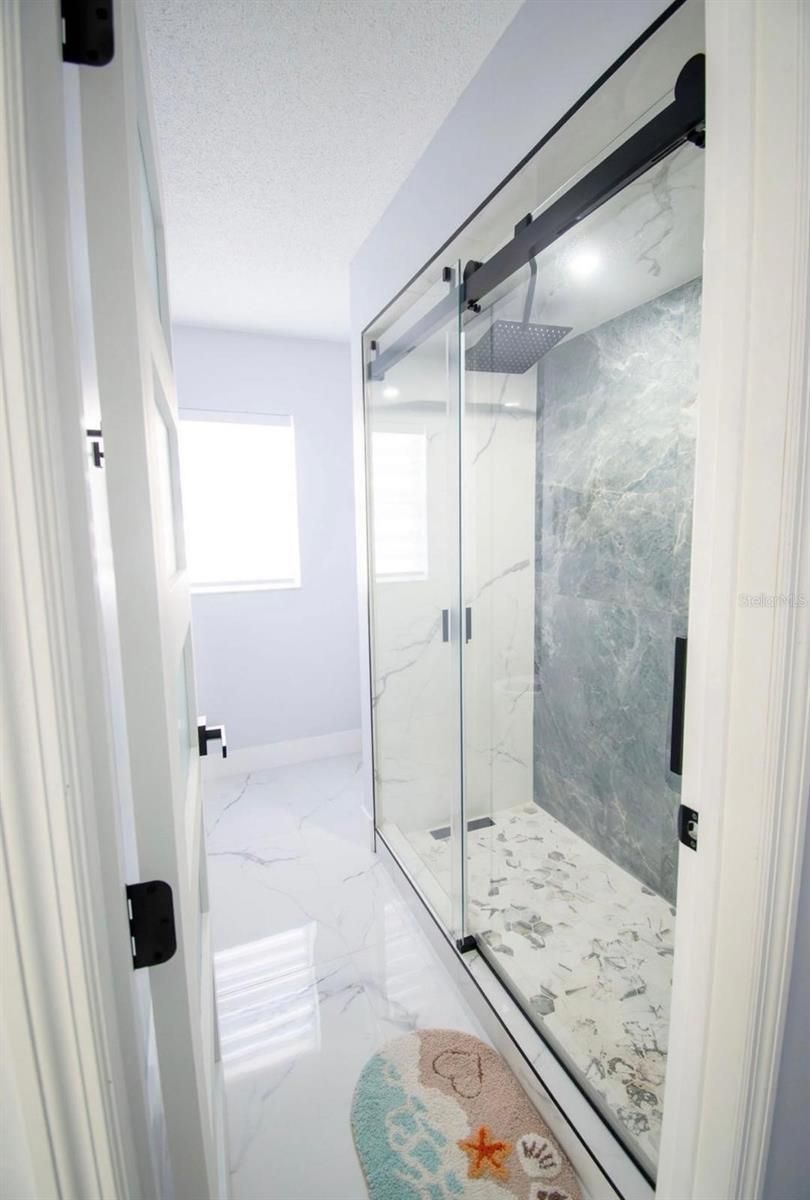 Pasillo de baño luminoso con ducha de cristal y suelo de baldosas de mármol blanco.