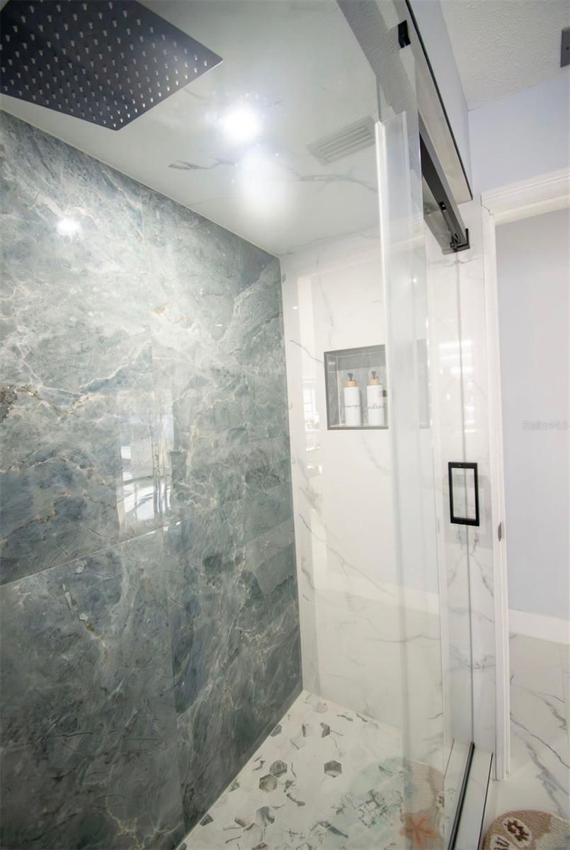 Ducha moderna con puerta de cristal, paredes revestidas de mármol y suelo de baldosas blancas.