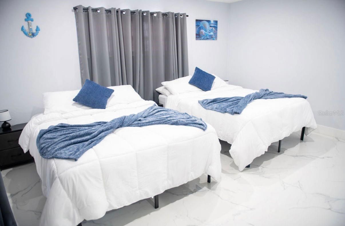 Dos camas individuales con ropa de cama azul en una habitación blanca luminosa con cortinas grises.