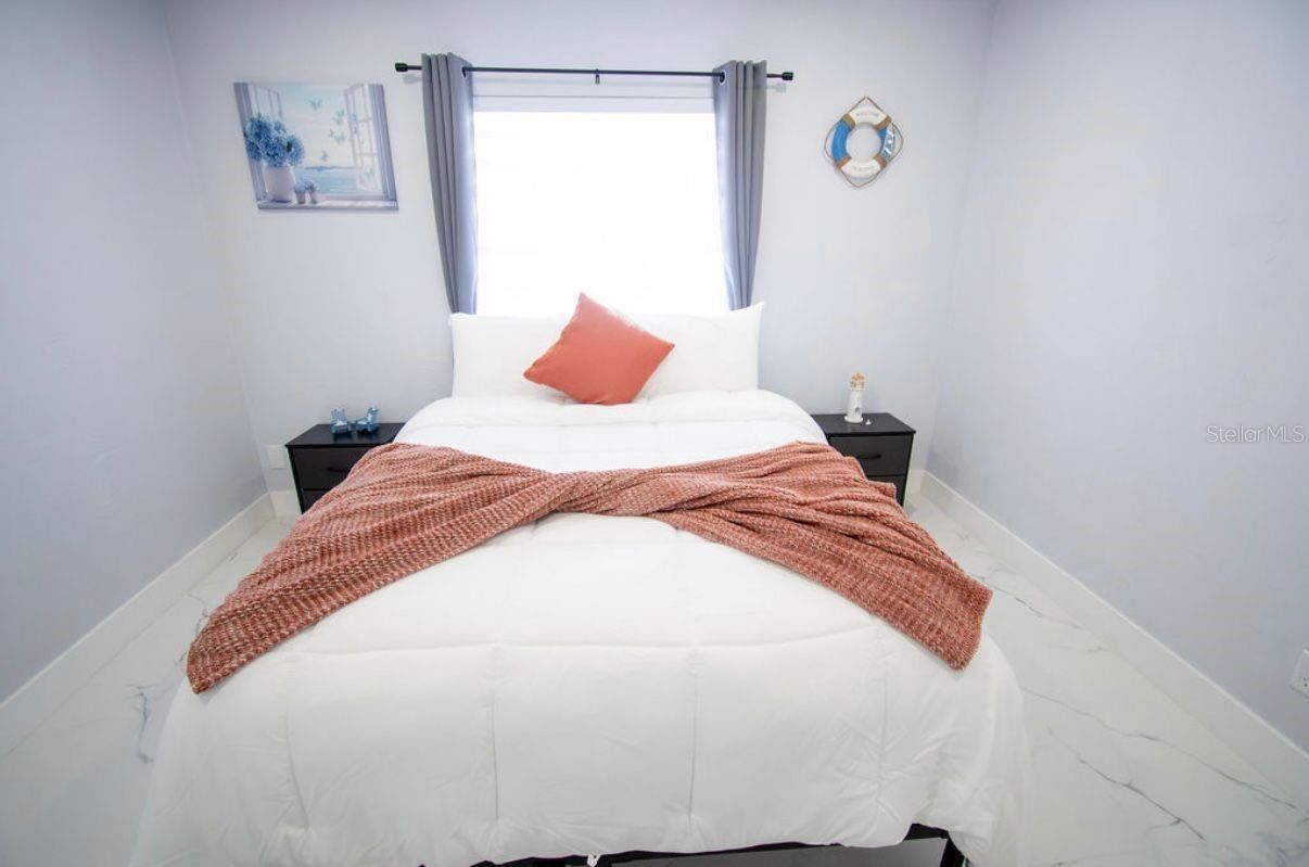 Cama blanca con manta color óxido y almohada color coral en un dormitorio minimalista y luminoso con ventana.