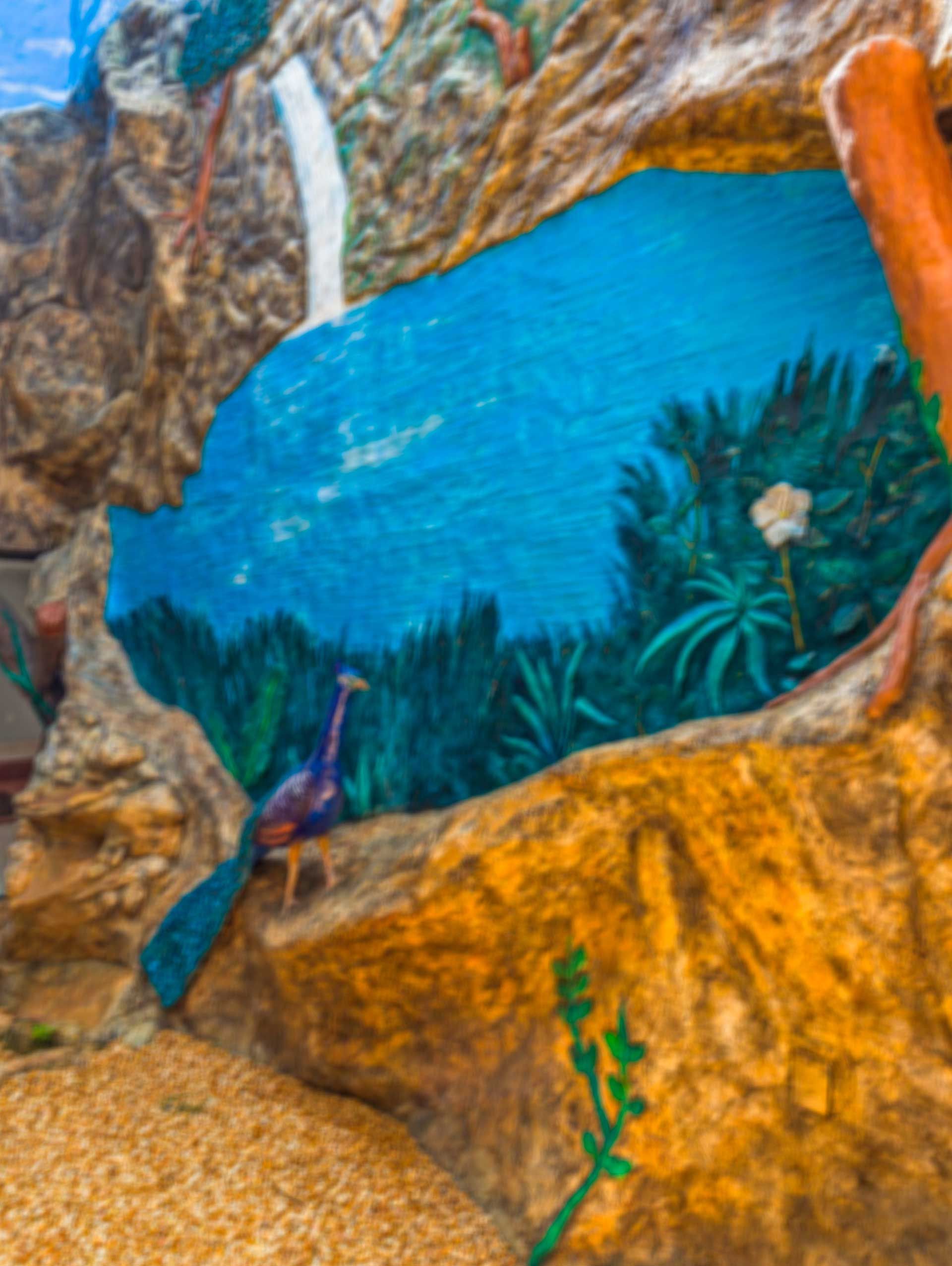Mural colorido de un pavo real en una cornisa rocosa, con vista a una escena de lago azul pintado con cascada y follaje.