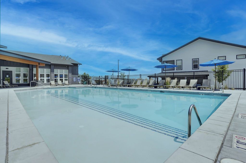 Outdoor community pool with lounge chairs and blue umbrellas, bordered by buildings.