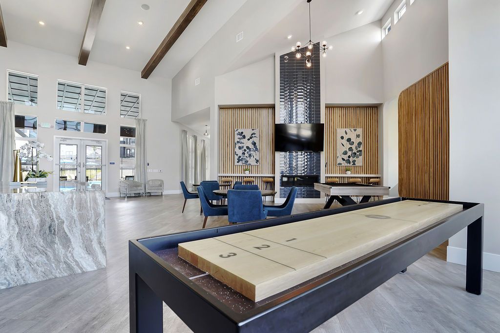 Modern resident lounge with a shuffleboard table, blue chairs, and a wall TV.