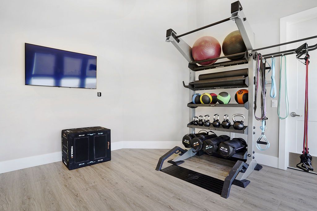 Indoor apartment gym with a weight rack, kettlebells, medicine balls, resistance bands, and a wall-mounted TV.