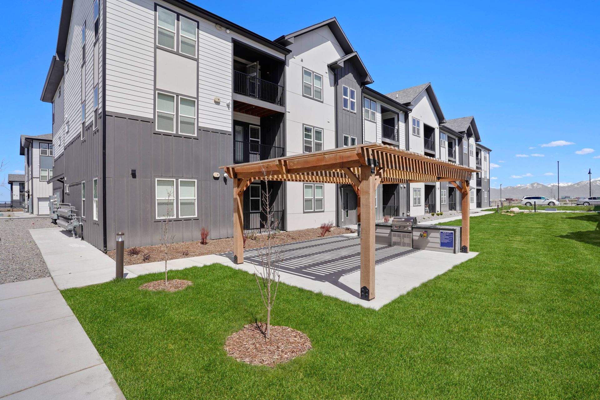 Exterior view of a modern apartment building with a communal grill beneath a wooden pergola.