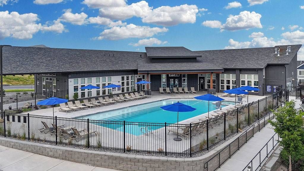 Outdoor community pool with lounge chairs, blue umbrellas, and a surrounding fence.