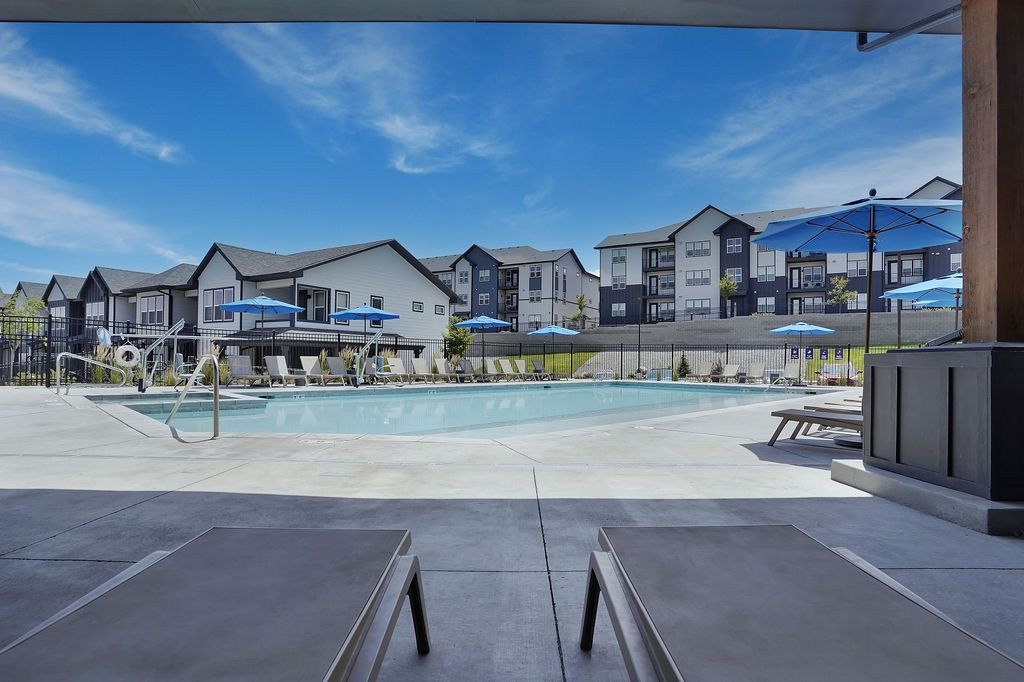 Outdoor pool area with lounge chairs and blue umbrellas beside modern apartment buildings.