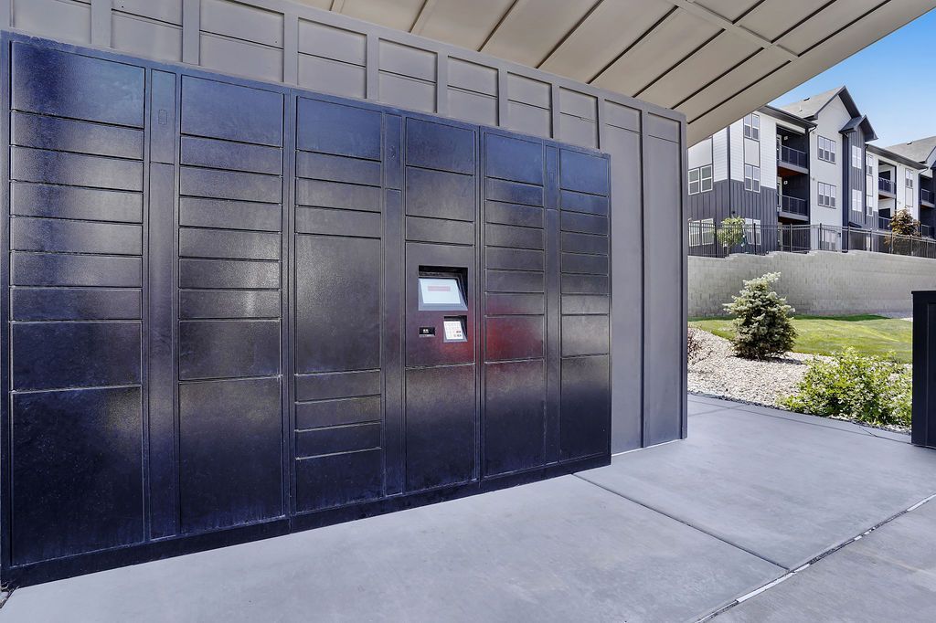 Black parcel/mail lockers under a covered structure with apartment buildings in the background.