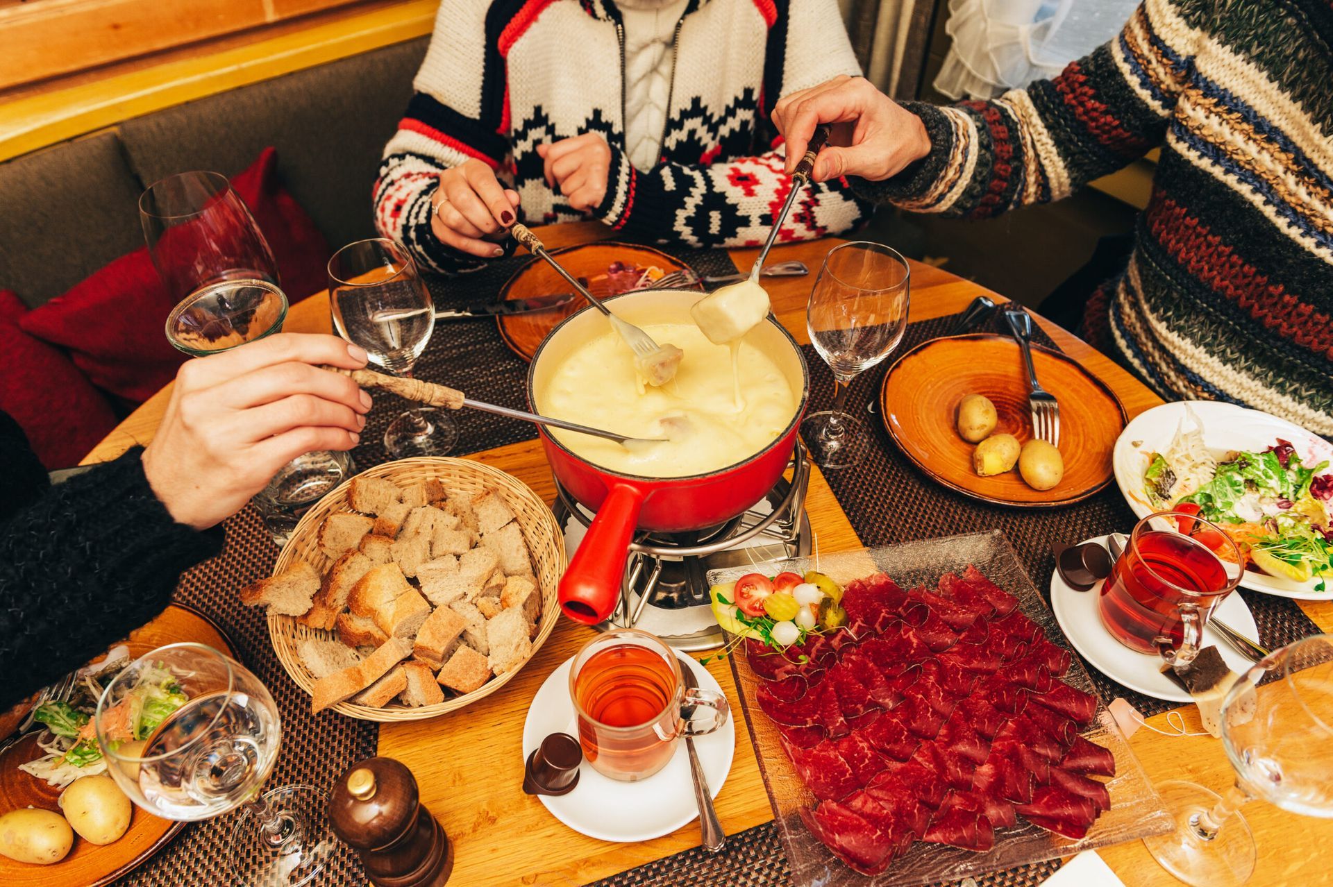 Hands dipping bread into a red fondue pot on a table with salads and potatoes