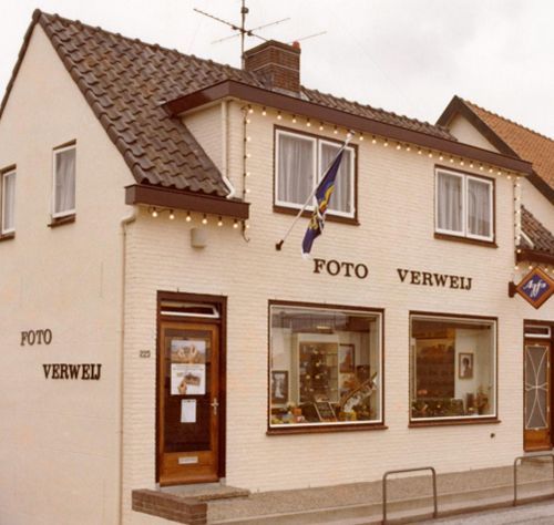 Corner photo shop with cream facade, brown roof, and a flag; sign reads “FOTO VERWEIJ.”