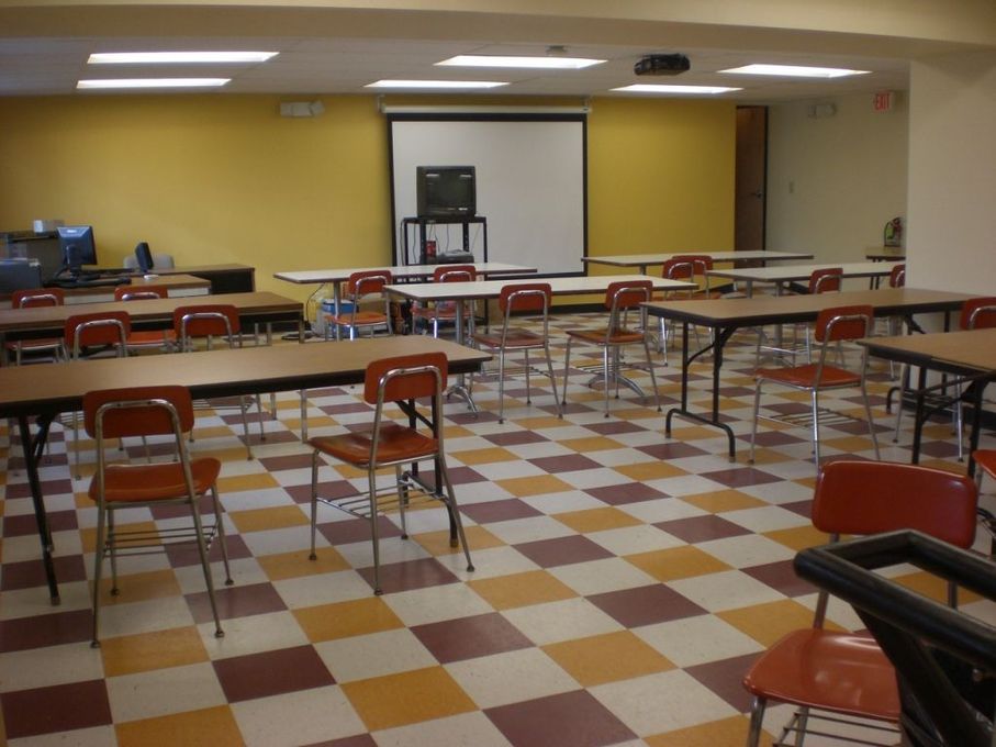 Classroom with brown and white checkered floor, yellow wall, tables, chairs, projector screen.