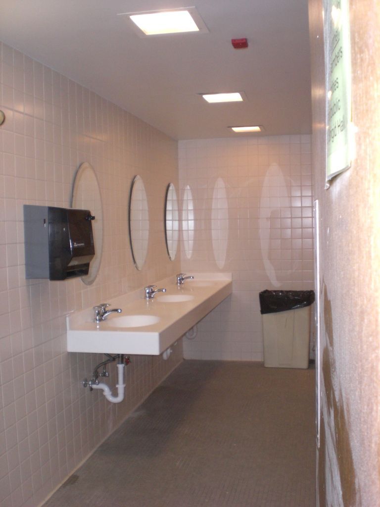 Public restroom with sinks, mirrors, and a trash can. White tile walls, gray floor, overhead lighting.