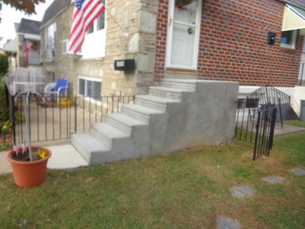 Concrete steps leading up to a brick and stone house. A small black fence and potted plant sit in the foreground.