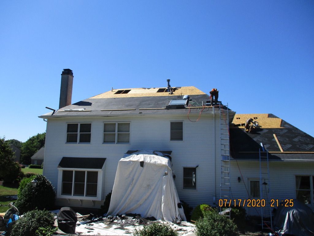 Roof of a white house being replaced on a sunny day. Workers on roof, tarp covers below, ladder present.