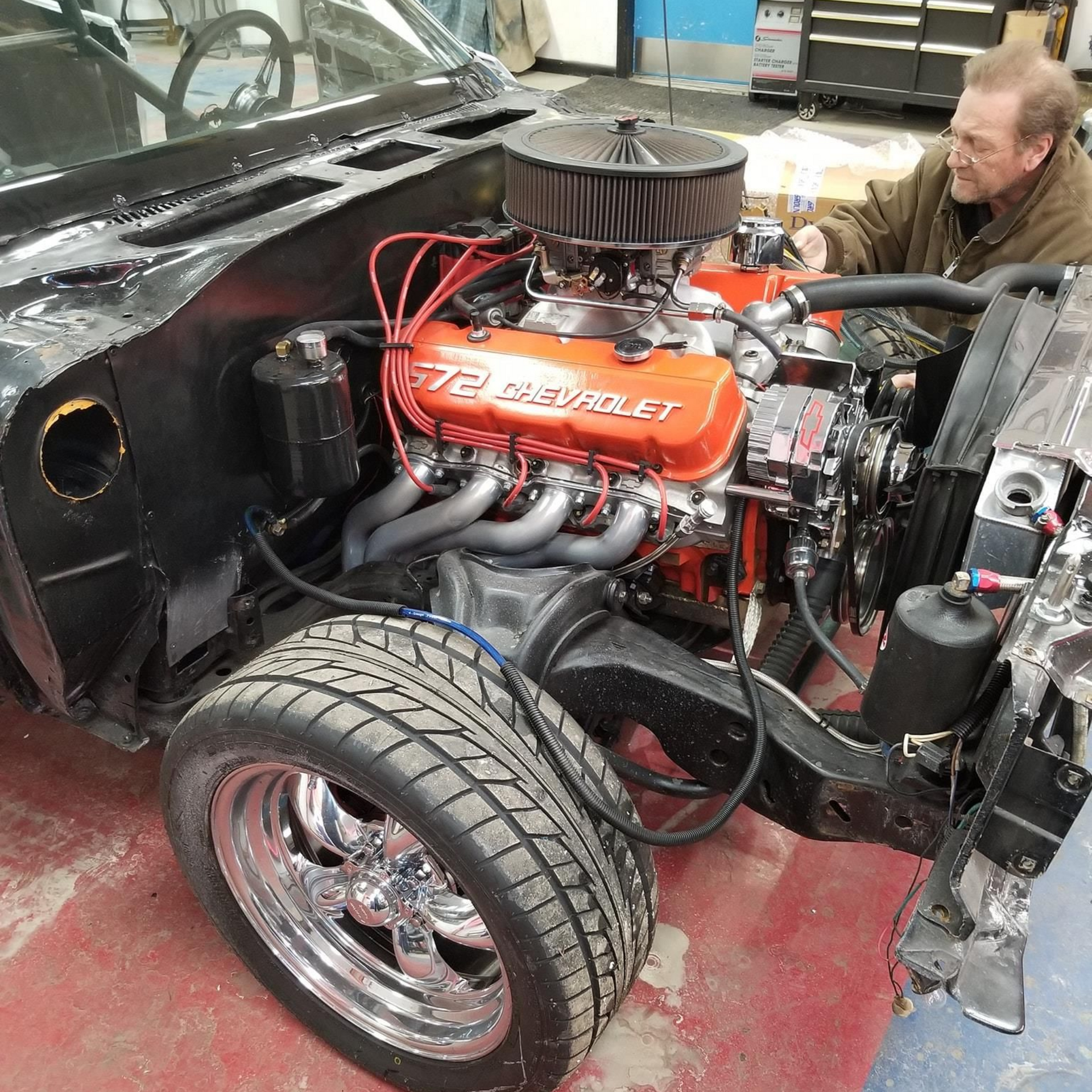 Black car engine bay with a large orange V8 Chevy engine, man working.