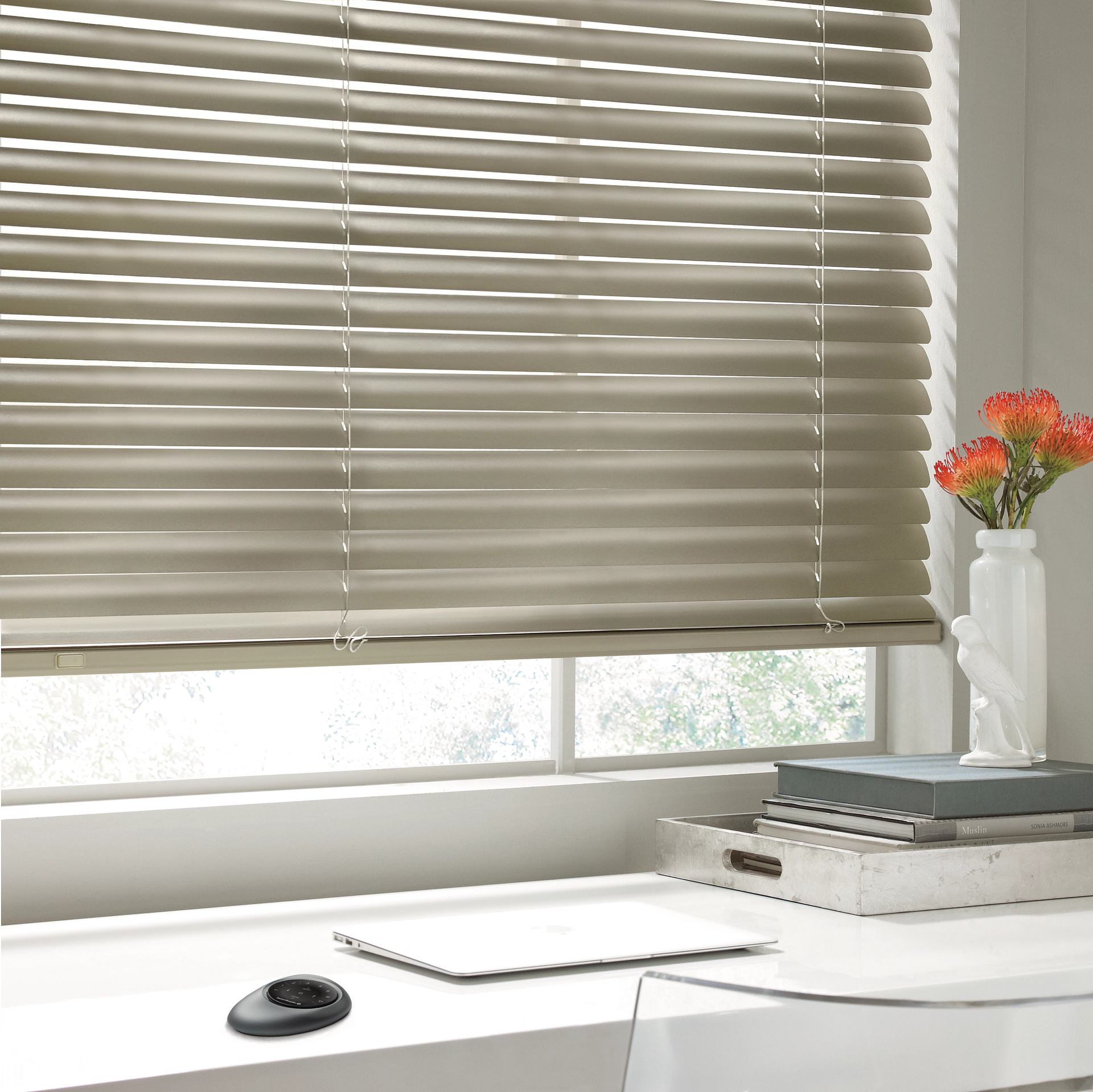 Bright desk by window with closed blinds, laptop, and vase of orange flowers on white shelf.