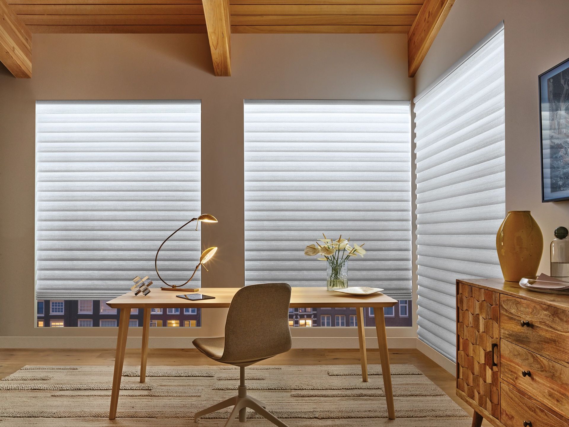 Modern home office with wooden desk, chair, lamp, flowers, and large blinds by a corner window