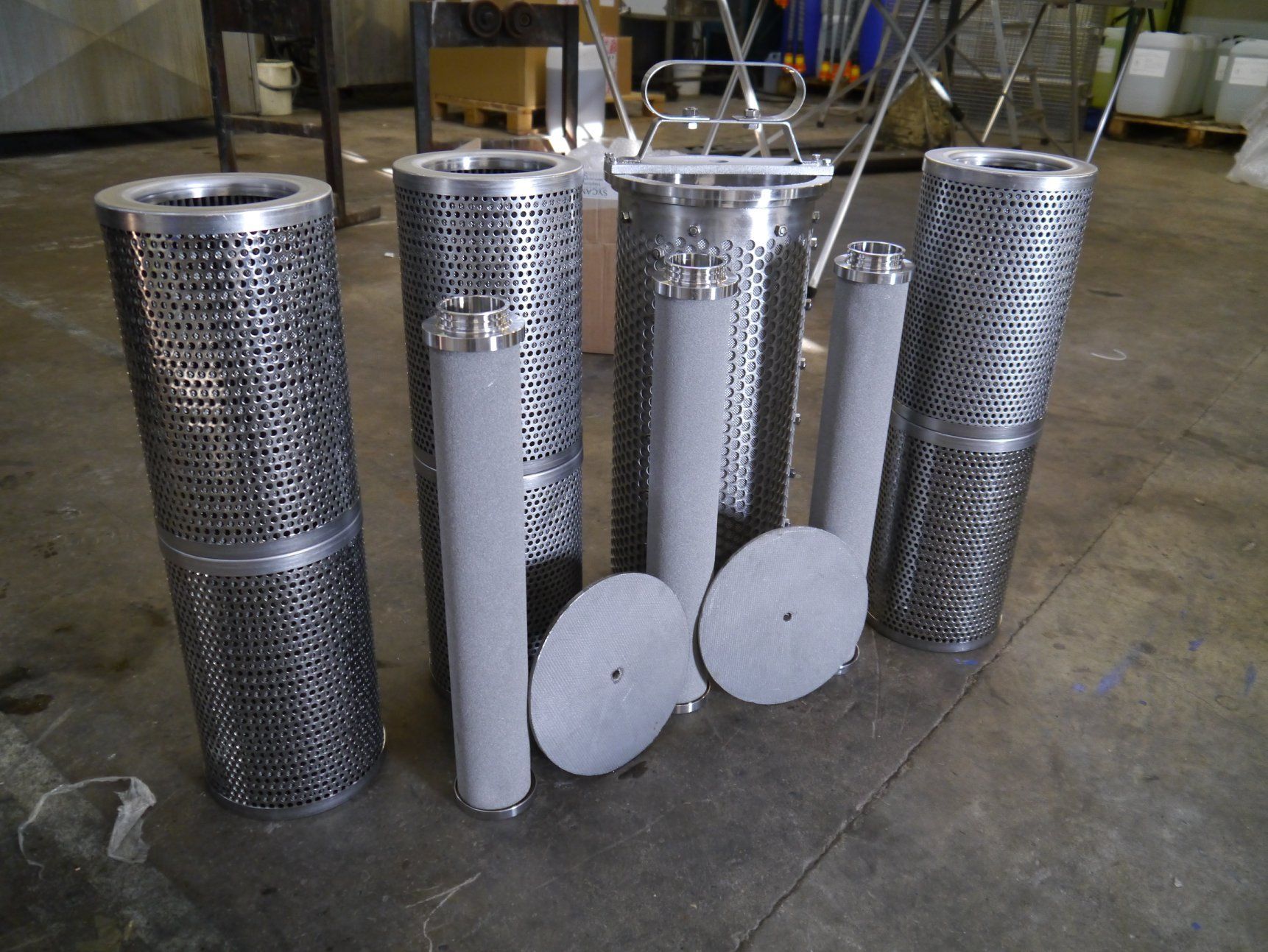 Keli metaliniai filtrai ir filtro elementai ant betoninių grindų; pramoninė aplinka.