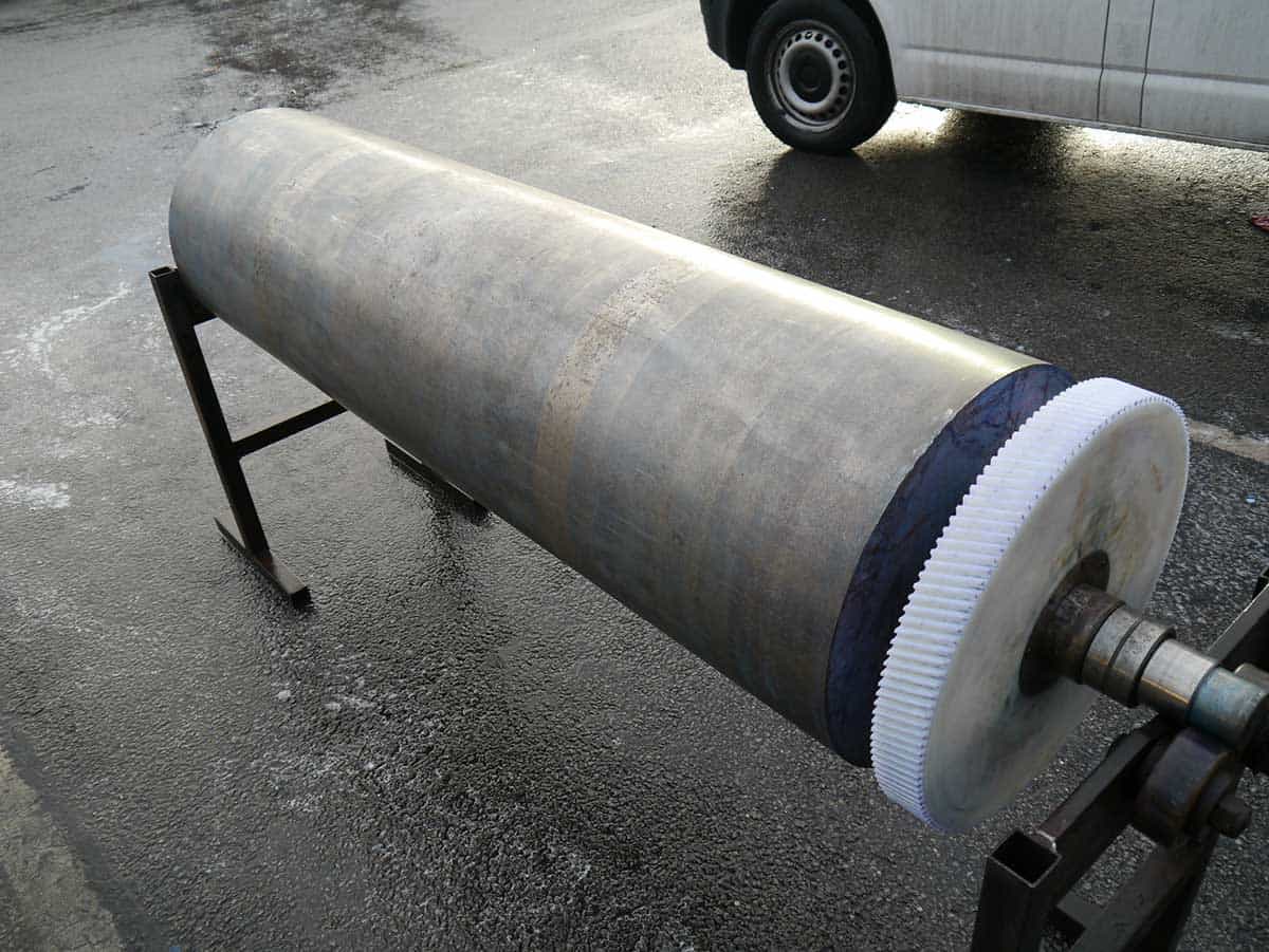 Didelis pilkas pramoninis volas su dantytu galu, pritvirtintas prie juodų metalinių atramų, lauke.