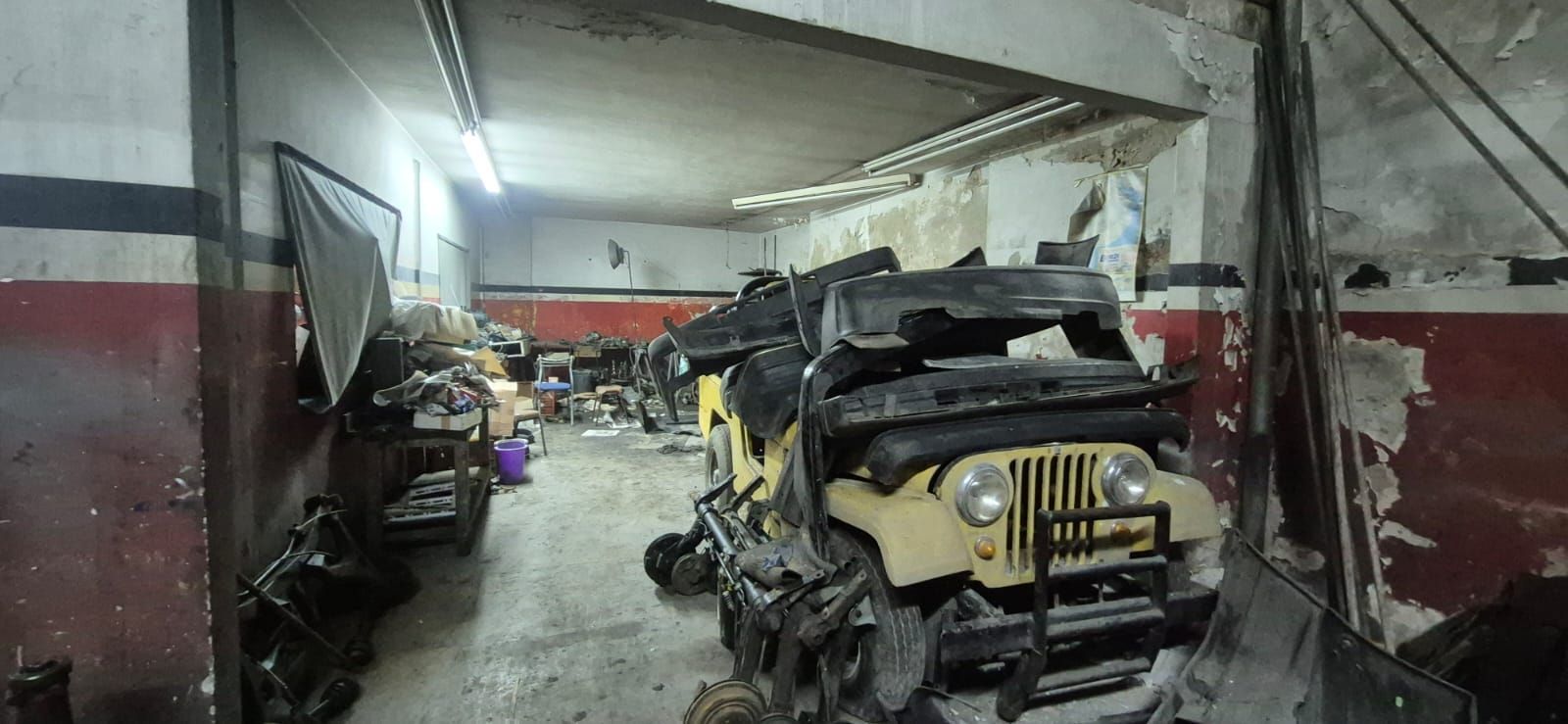 Un garaje desordenado con varios jeeps desmontados, paredes rojas y blancas y una luz fluorescente.