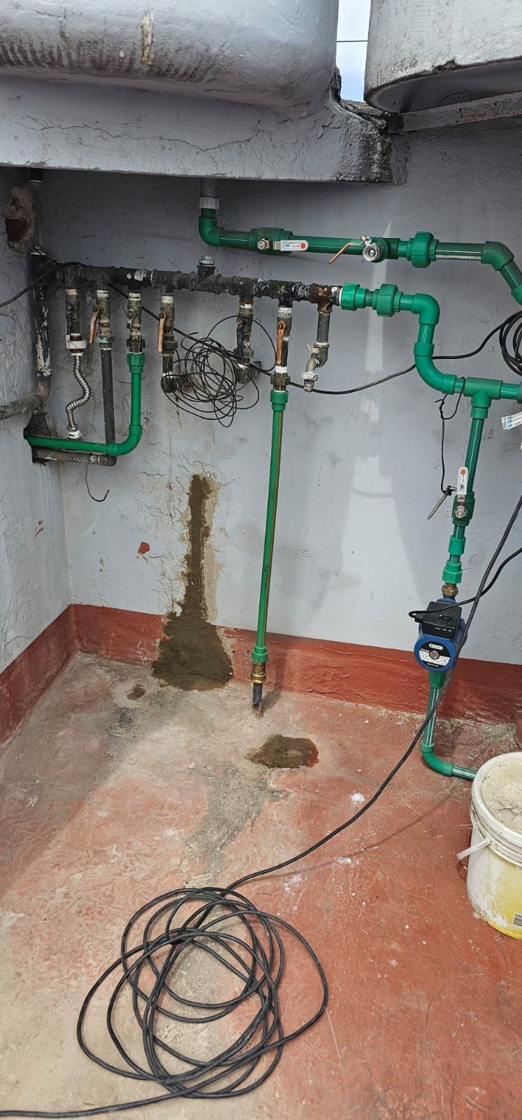 Tuberías de plomería sobre un piso de concreto, con una mancha marrón en la pared y una bobina de cable en el piso.