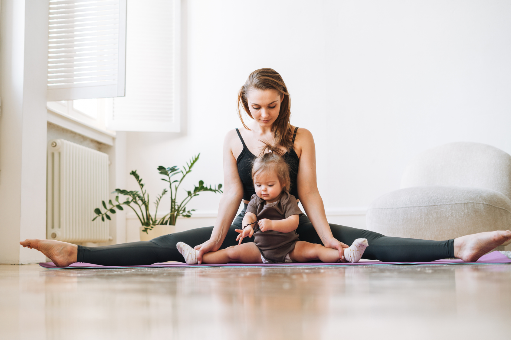 Mother in a wide-legged seated stretch with baby sitting between her legs during Mommy & Me Pilates.