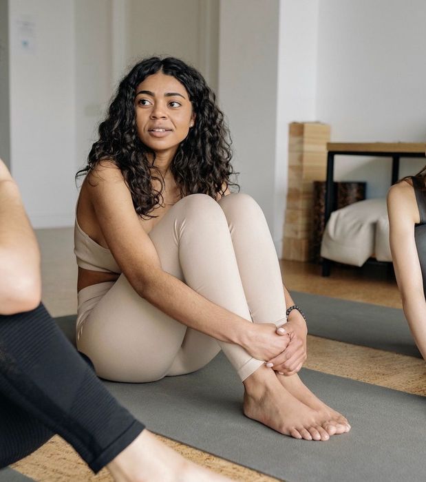 Woman sitting on a mat with arms wrapped around knees, listening attentively during a Pilates class.