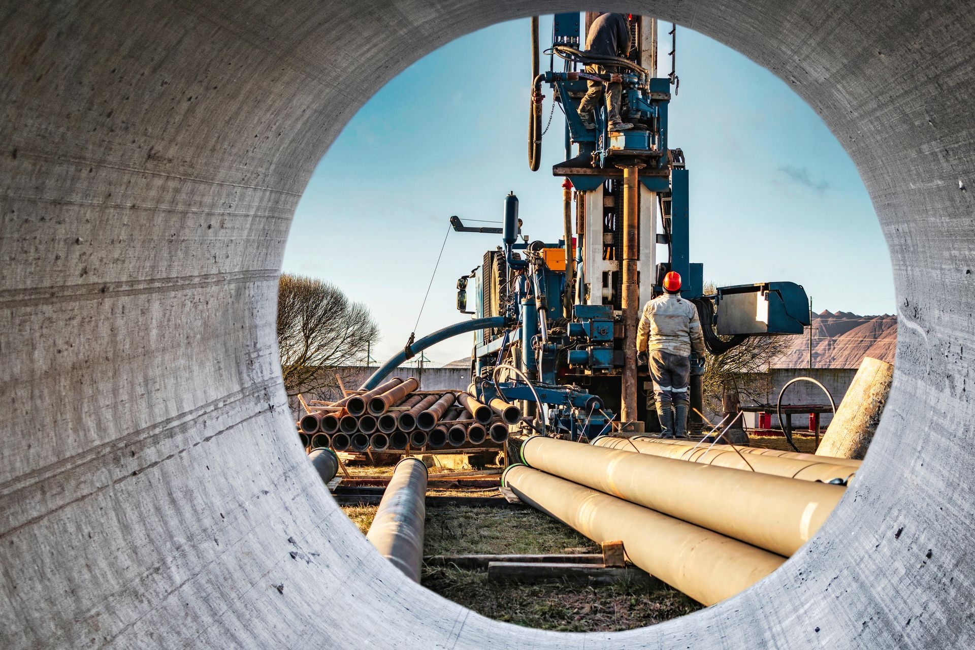 Workers at a pipeline construction site framed through a large concrete pipe opening. Workers at a pipeline construction site framed through a large concrete pipe opening.