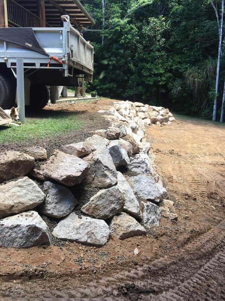 a Rock Retaining Wall Being Built on a Dirt Driveway, Truck Parked Behind It — Macca's Dynamic Earthworks in Tolga, QLD