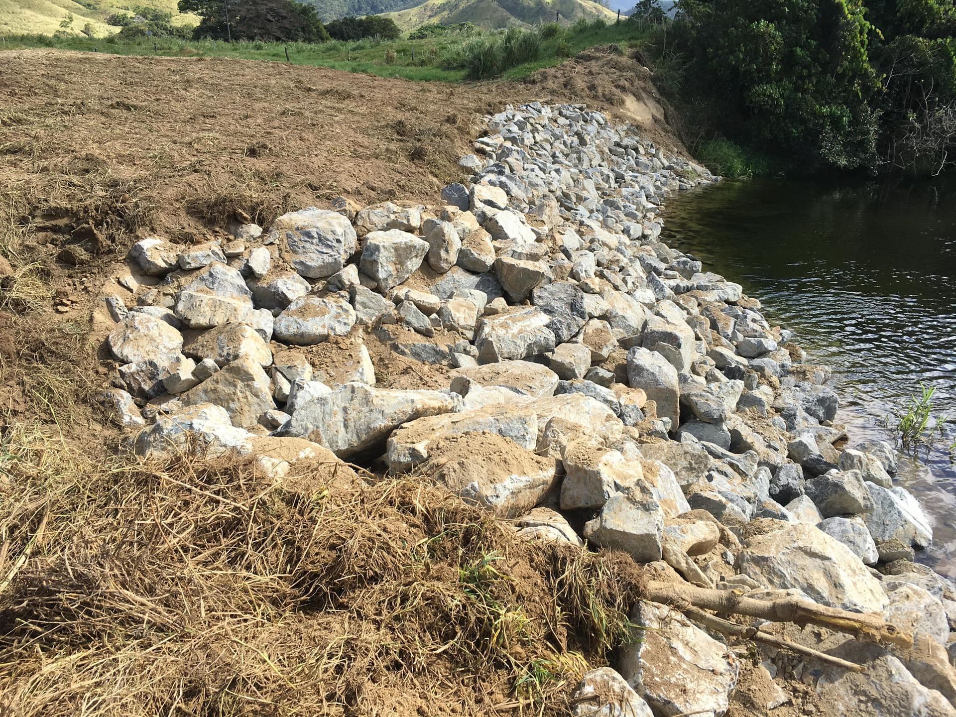 A Rocky Shoreline with A Pile of Brown Debris, Next to A Calm River — Macca's Dynamic Earthworks in Tolga, QLD