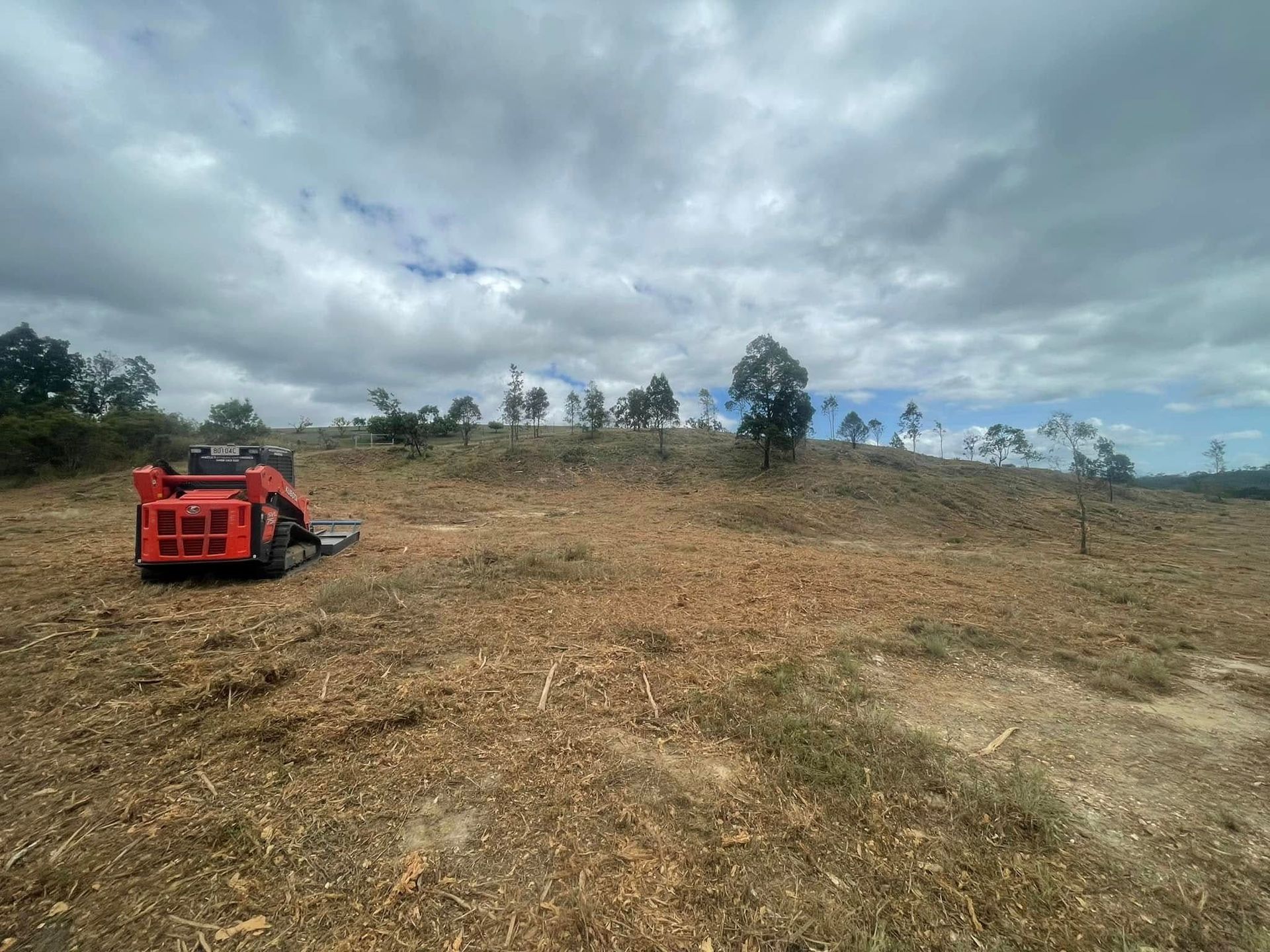 Orange Skid Steer Clearing a Field Under Cloudy Sky — Macca's Dynamic Earthworks in Tolga, QLD