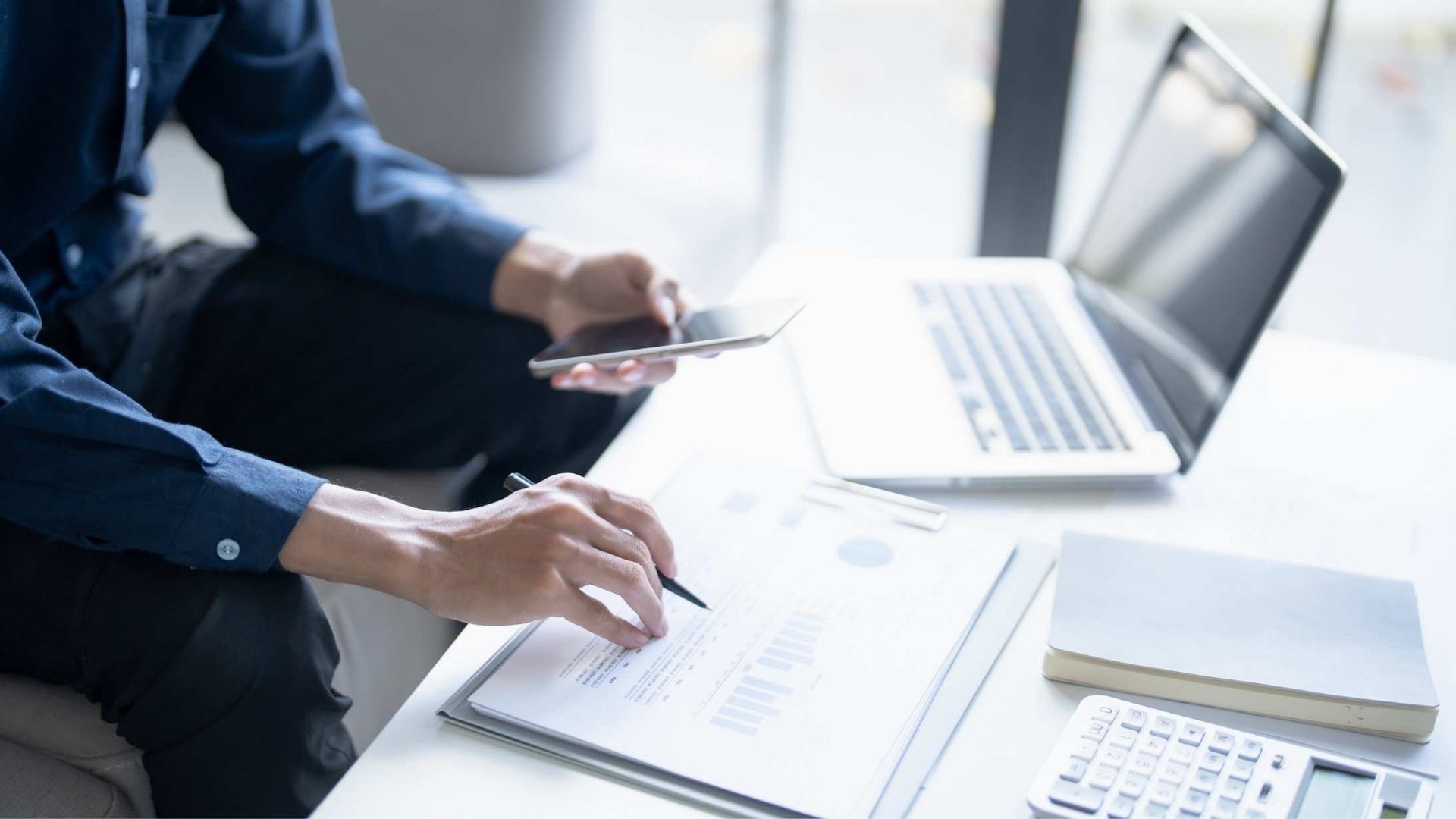 Person working on documents with a laptop, calculator, and phone on a table.