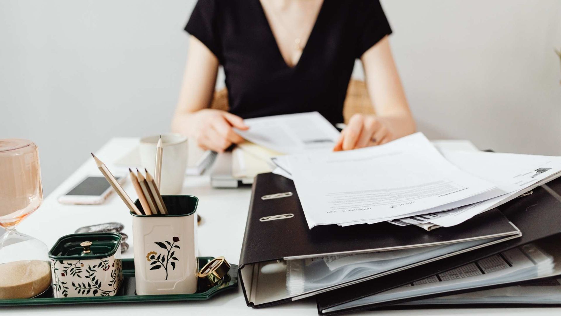 Woman at a white desk, sorting papers from binders. Office setting with office supplies.