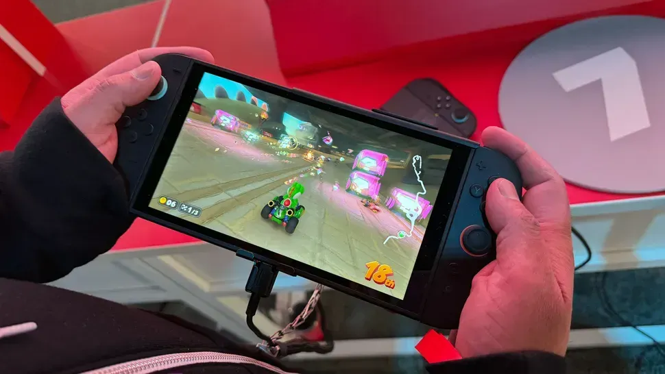 Person holding a black handheld gaming device playing Mario Kart. The screen shows a race.
