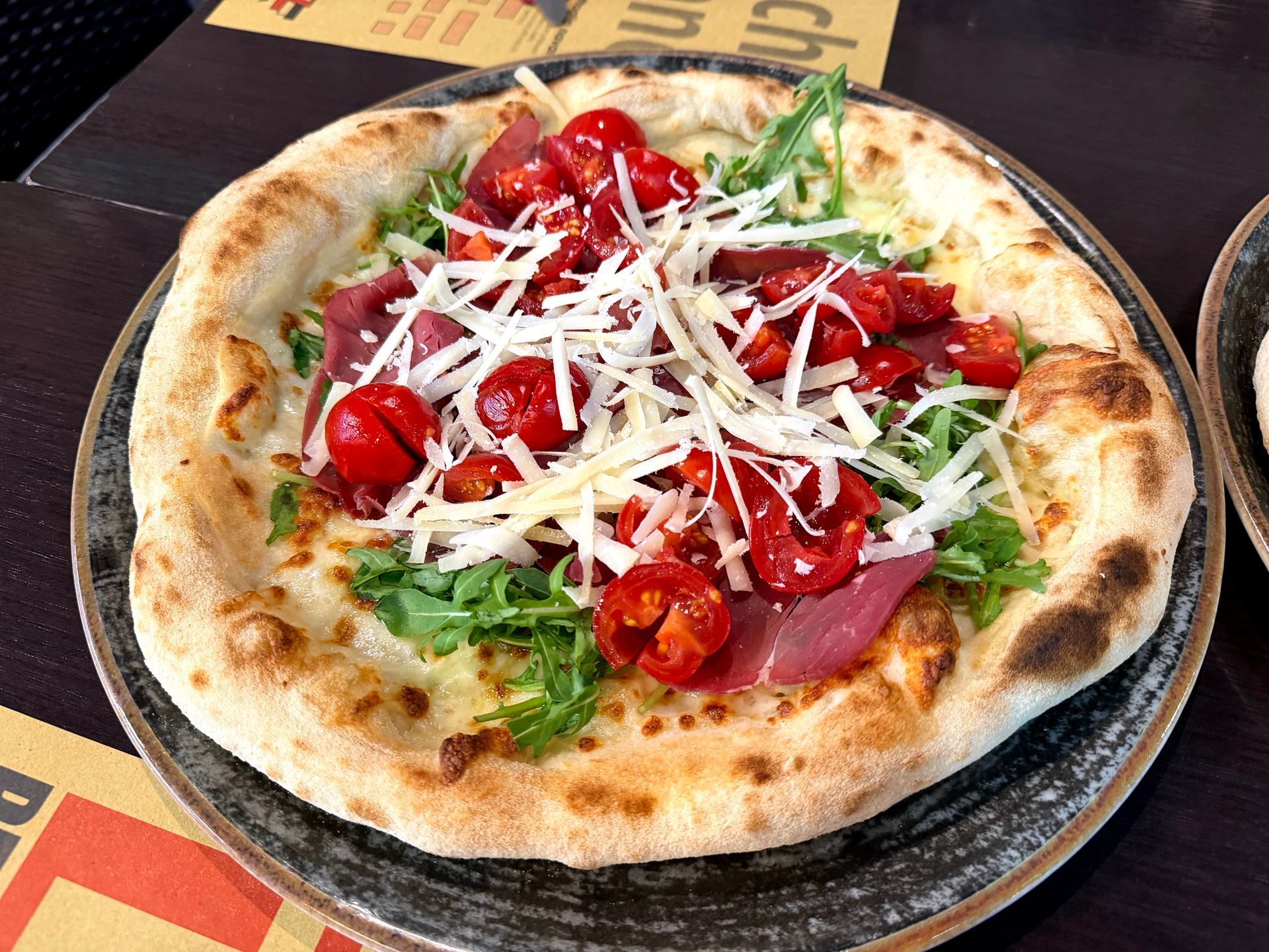 Pizza con pomodori, rucola, formaggio e prosciutto su un piatto scuro.