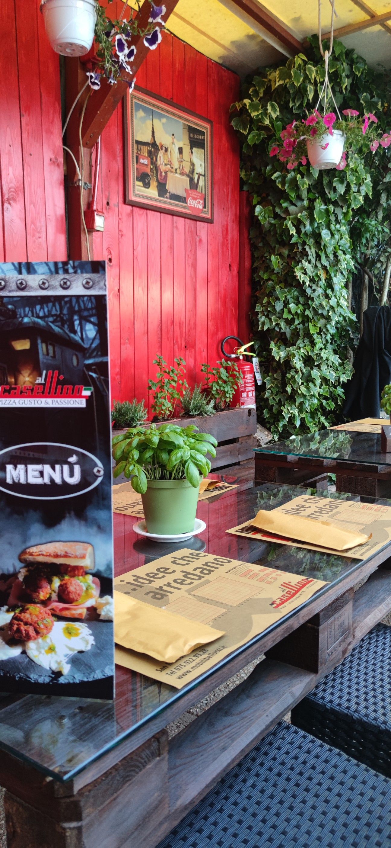 Patio del ristorante con muro rosso, piante verdi, tavolo in legno, menù e pianta di basilico.