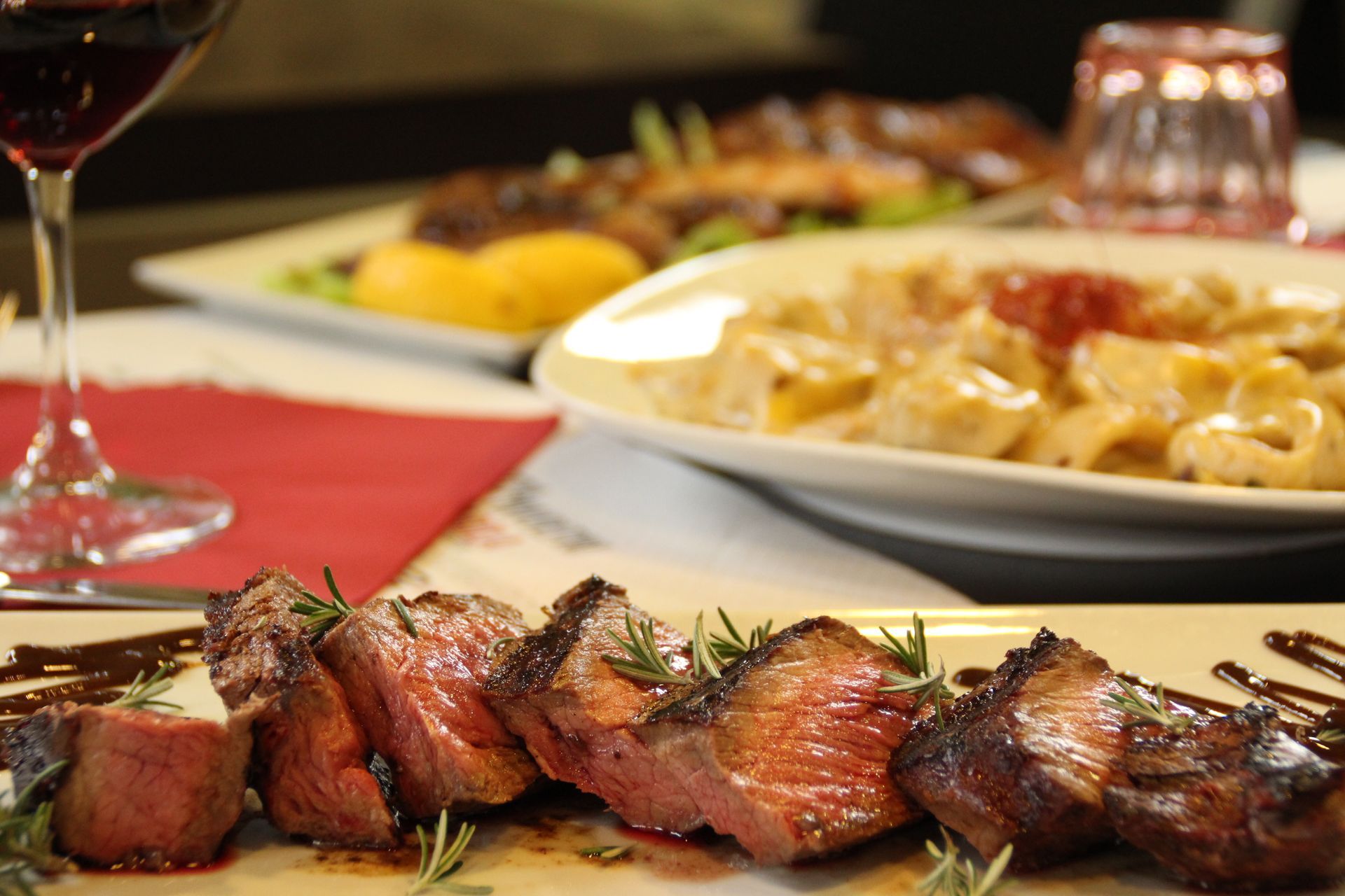 Bistecca tagliata al rosmarino su un piatto, altre pietanze e un bicchiere di vino sullo sfondo.