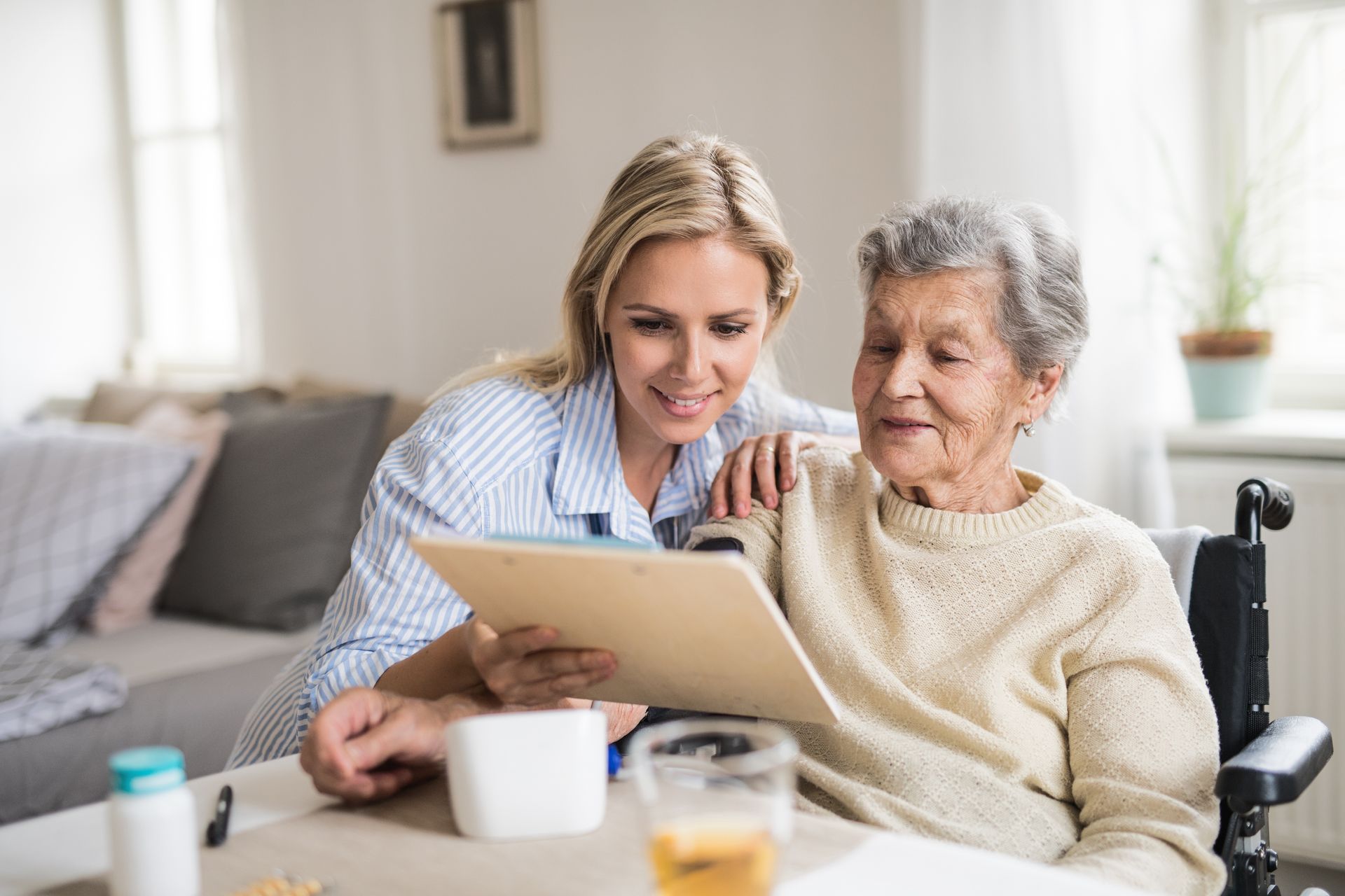 Woman assisting an older person in a wheelchair, both looking at a tablet indoors.